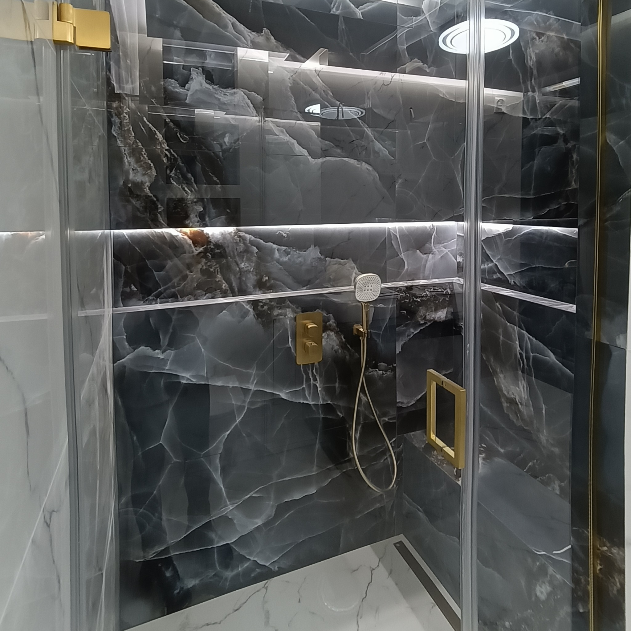 Kabina prysznicowa z czarnymi marmurowymi płytkami, złotymi detalami i oświetleniem LED. Nowoczesny design łazienki z eleganckimi akcentami.