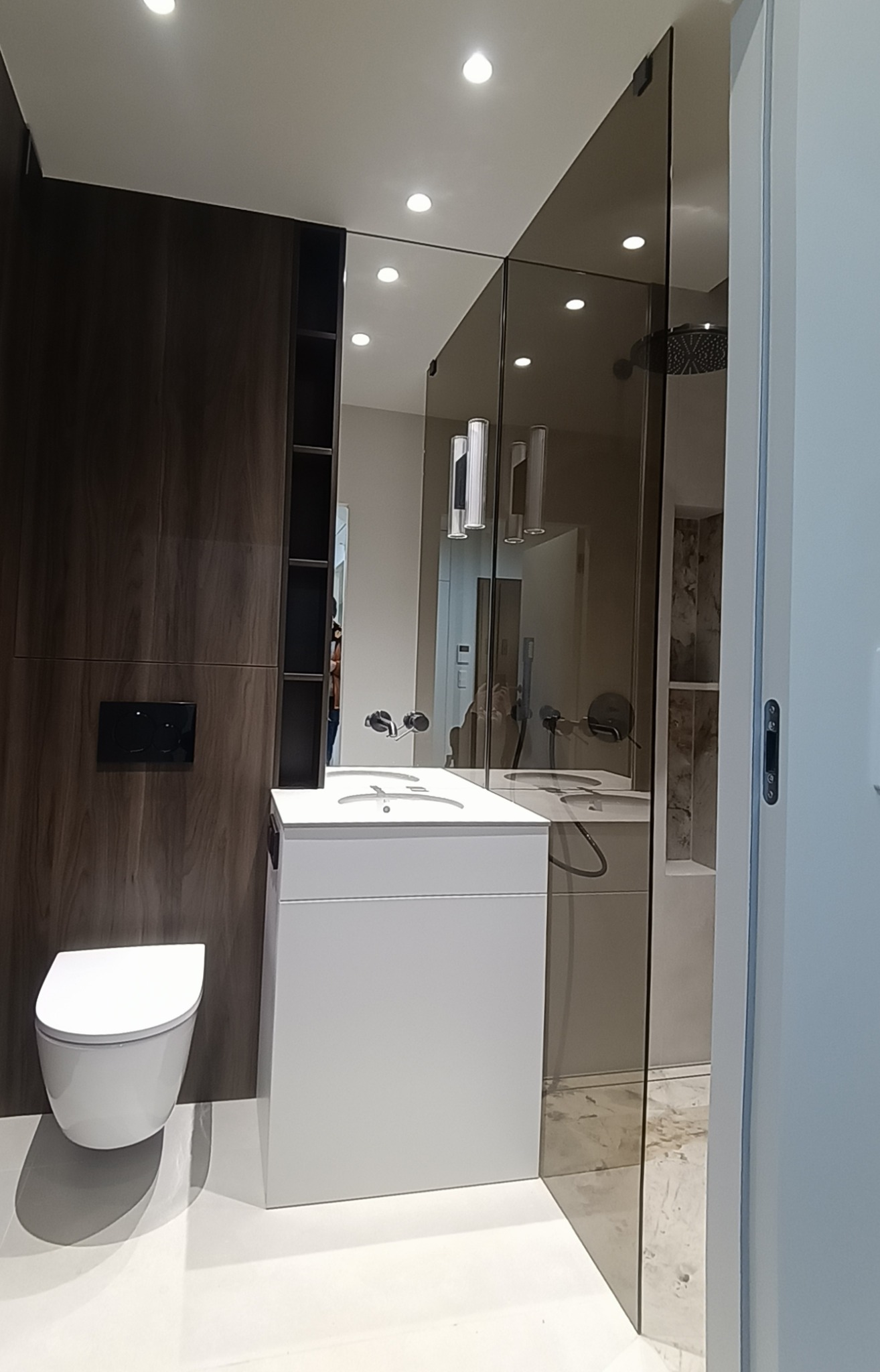 Nowoczesna łazienka z kabiną prysznicową ze szkła dymionego, białą umywalką i toaletą. Ciemne drewno na ścianie z wbudowanymi półkami. Oświetlenie punktowe.