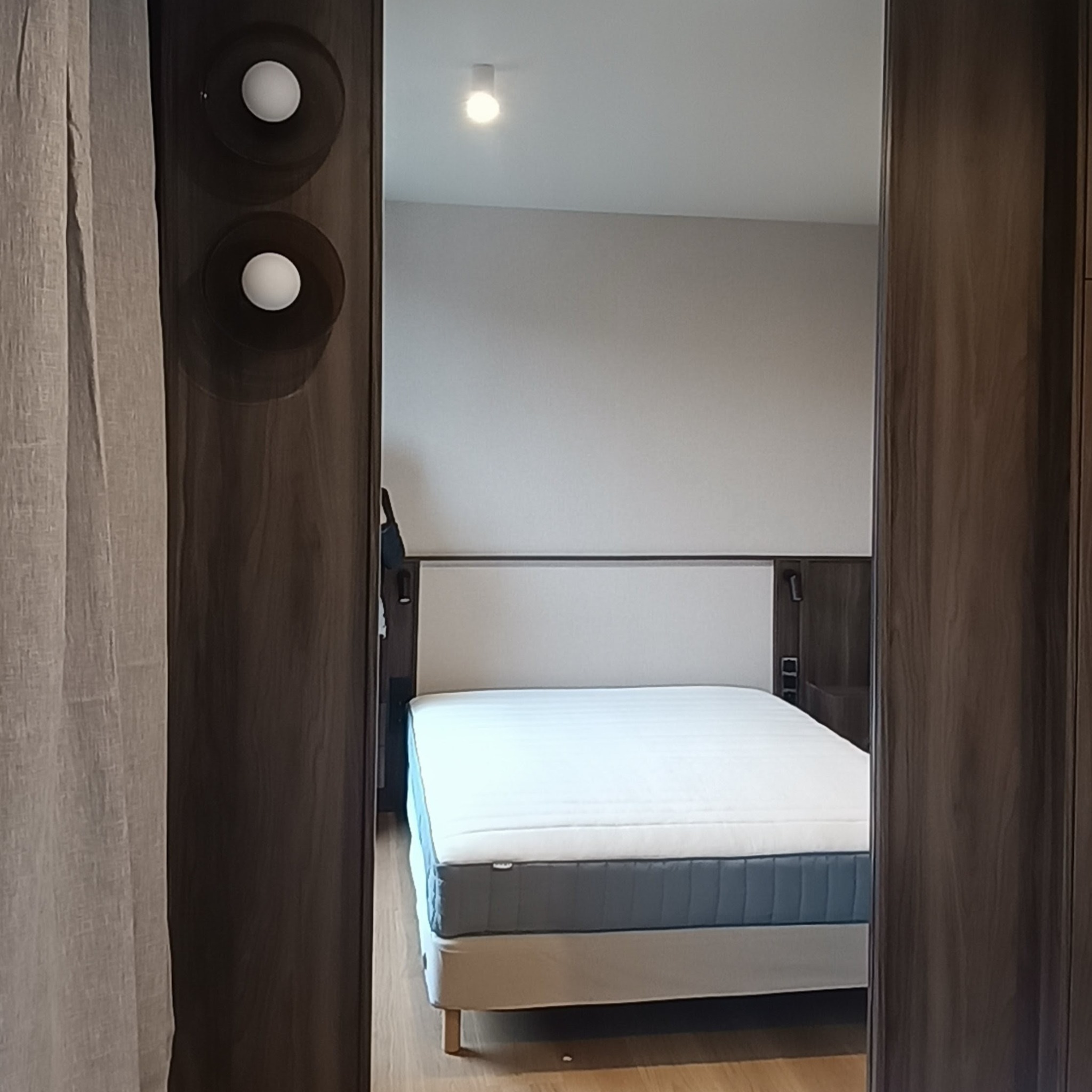 Nowoczesna sypialnia widziana przez otwarte, drewniane drzwi szafy. Widoczne łóżko z białą pościelą i szarym materacem, oświetlenie sufitowe i lampy na szafie.