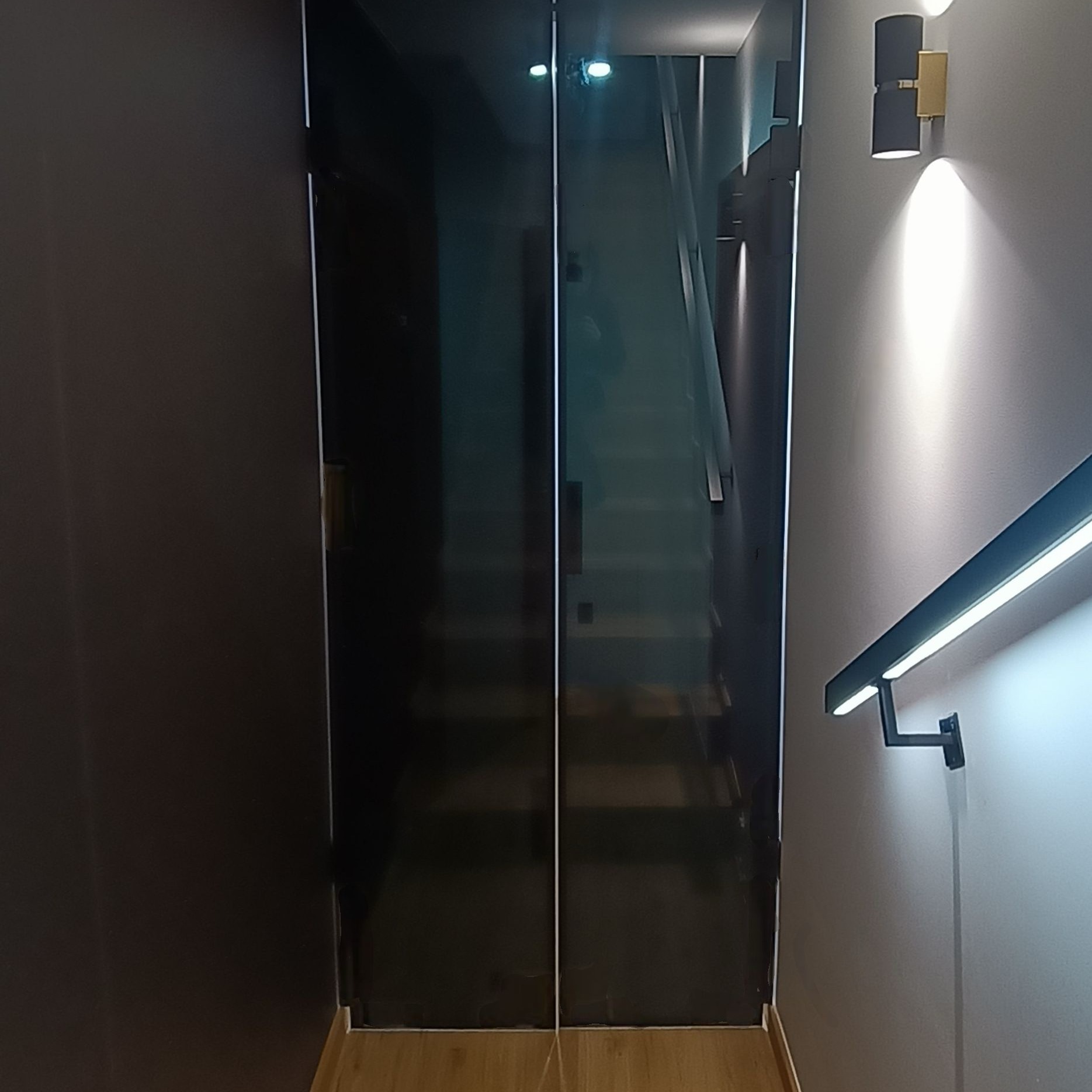 Szklane drzwi przesuwne z czarnymi okuciami, widok na schody. Nowoczesne oświetlenie ścienne. Minimalistyczny design.