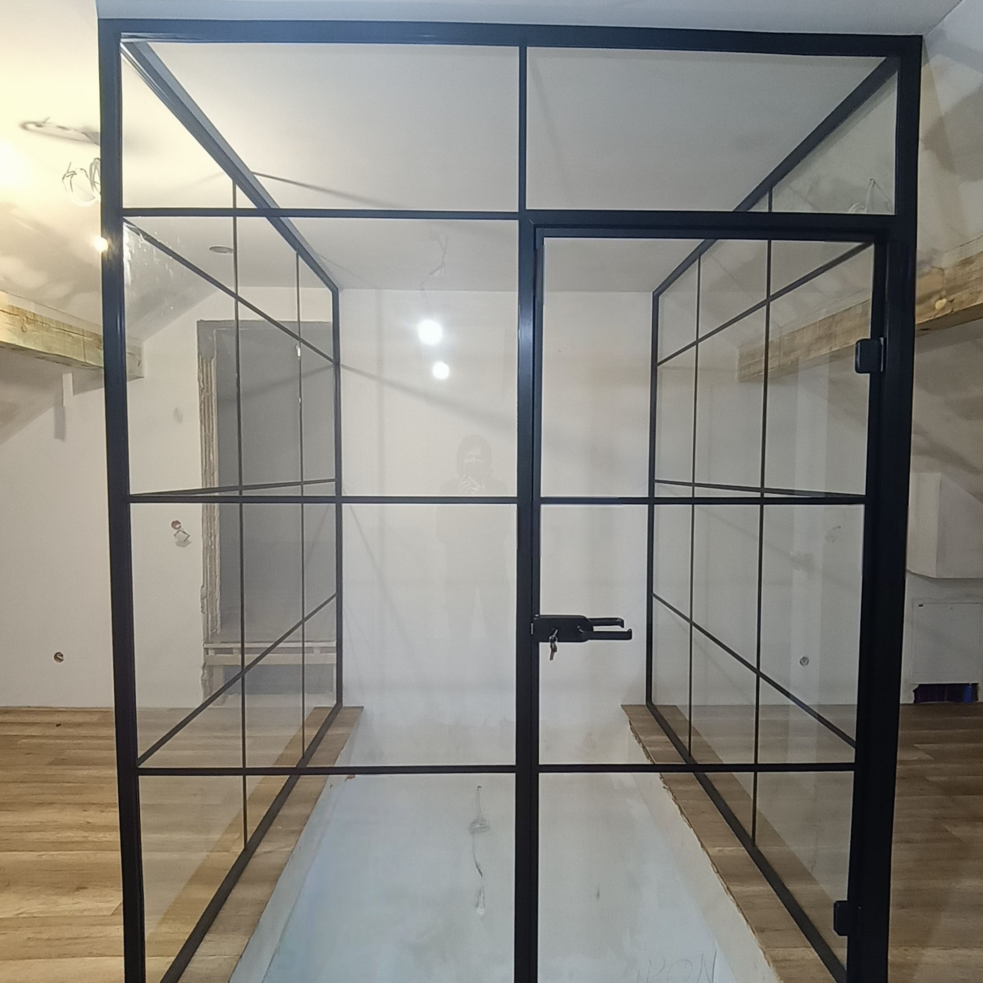 Nowoczesna, czarna, szklana ścianka działowa z drzwiami, oddzielająca przestrzeń w surowym wnętrzu. Widoczne drewniane elementy konstrukcyjne i podłoga.