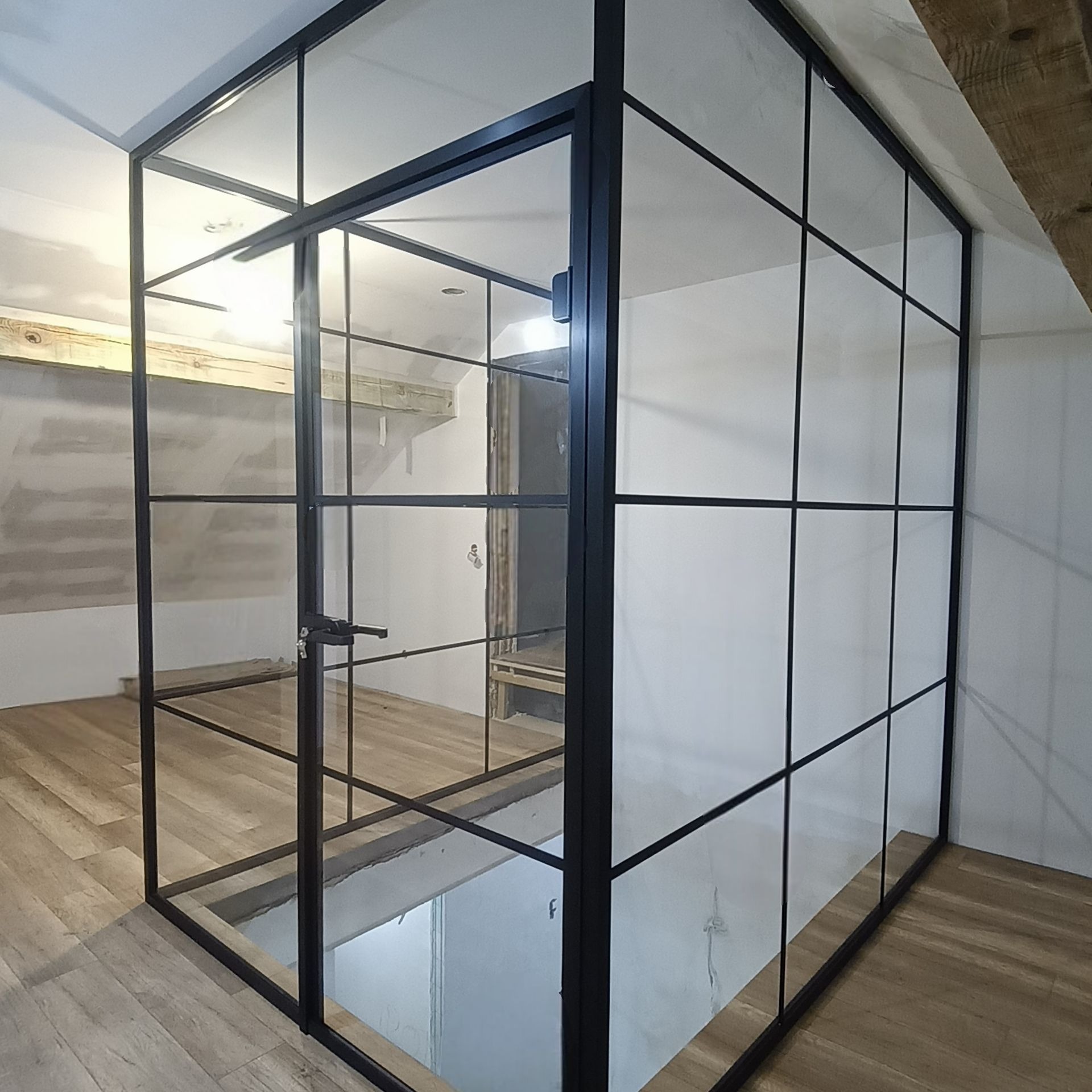 Nowoczesna, loftowa ścianka działowa ze szkła i czarnej stali, oddzielająca przestrzeń z drewnianą podłogą. Widoczna klamka i fragment surowego wykończenia.