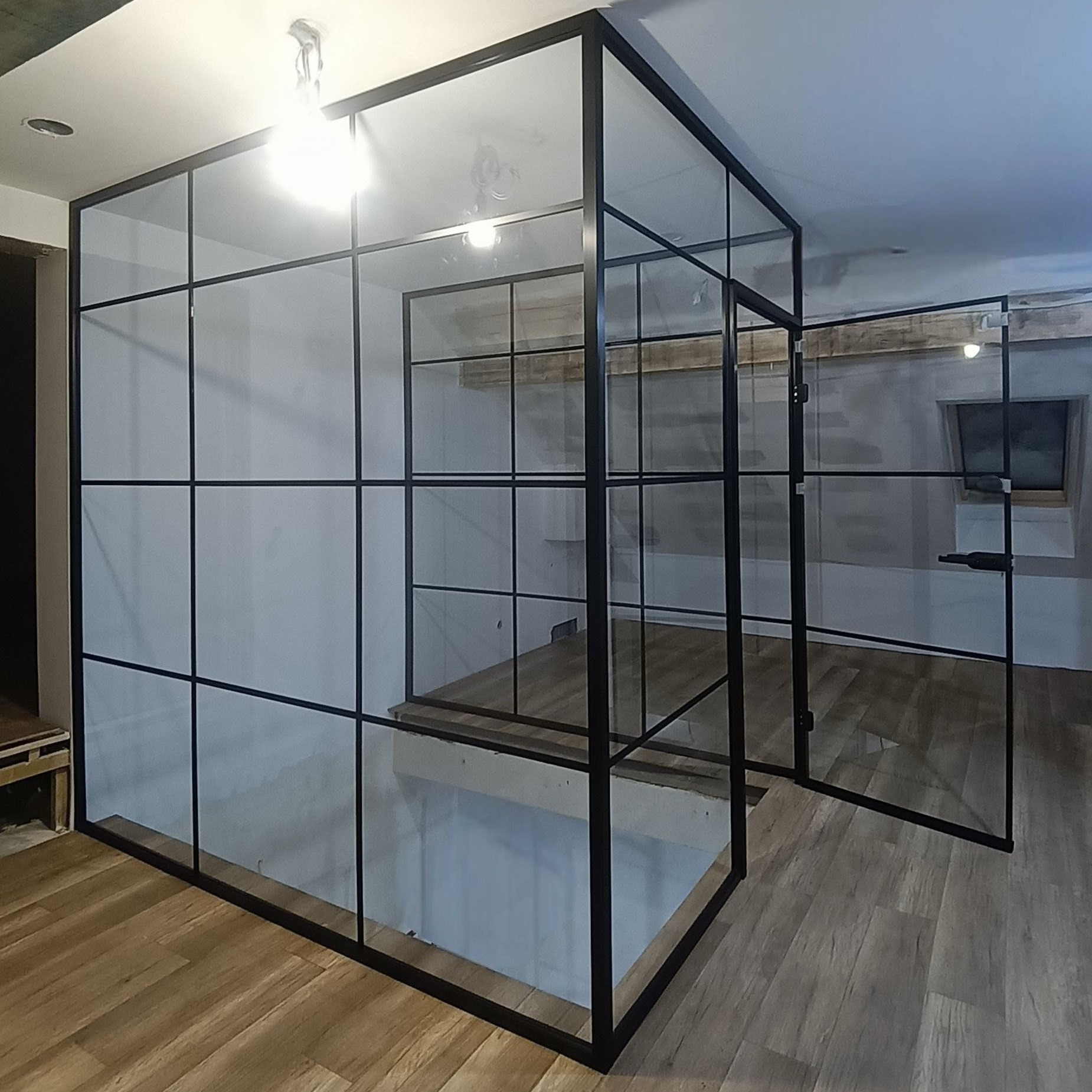 Nowoczesna, loftowa ścianka działowa ze szkła i czarnej stali, oddzielająca przestrzeń w jasnym wnętrzu. Widoczne drzwi i drewniana podłoga.