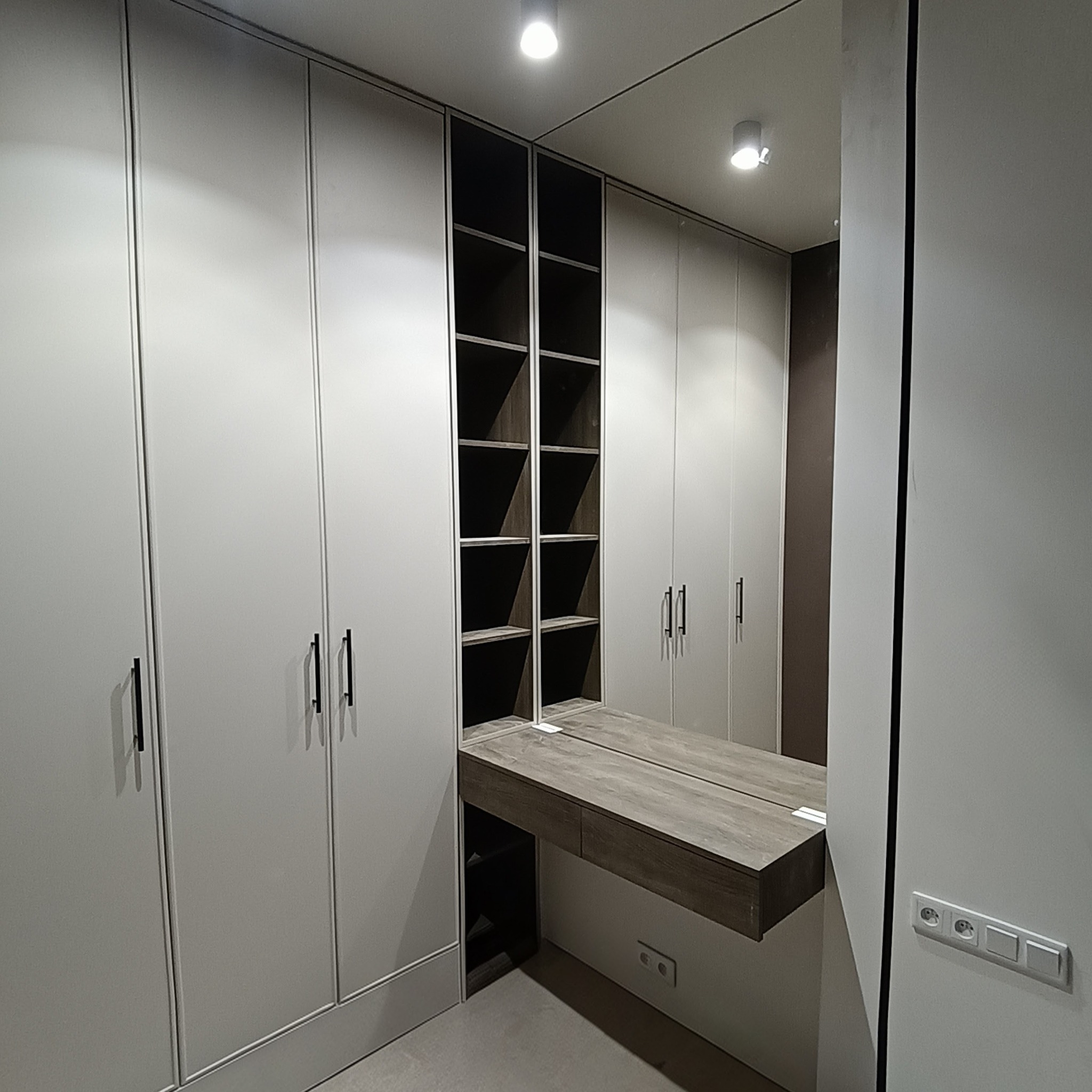 Nowoczesna garderoba z lustrem i półkami, jasne fronty szaf z minimalistycznymi uchwytami, oświetlenie punktowe, widok na wnętrze szafy z półkami.