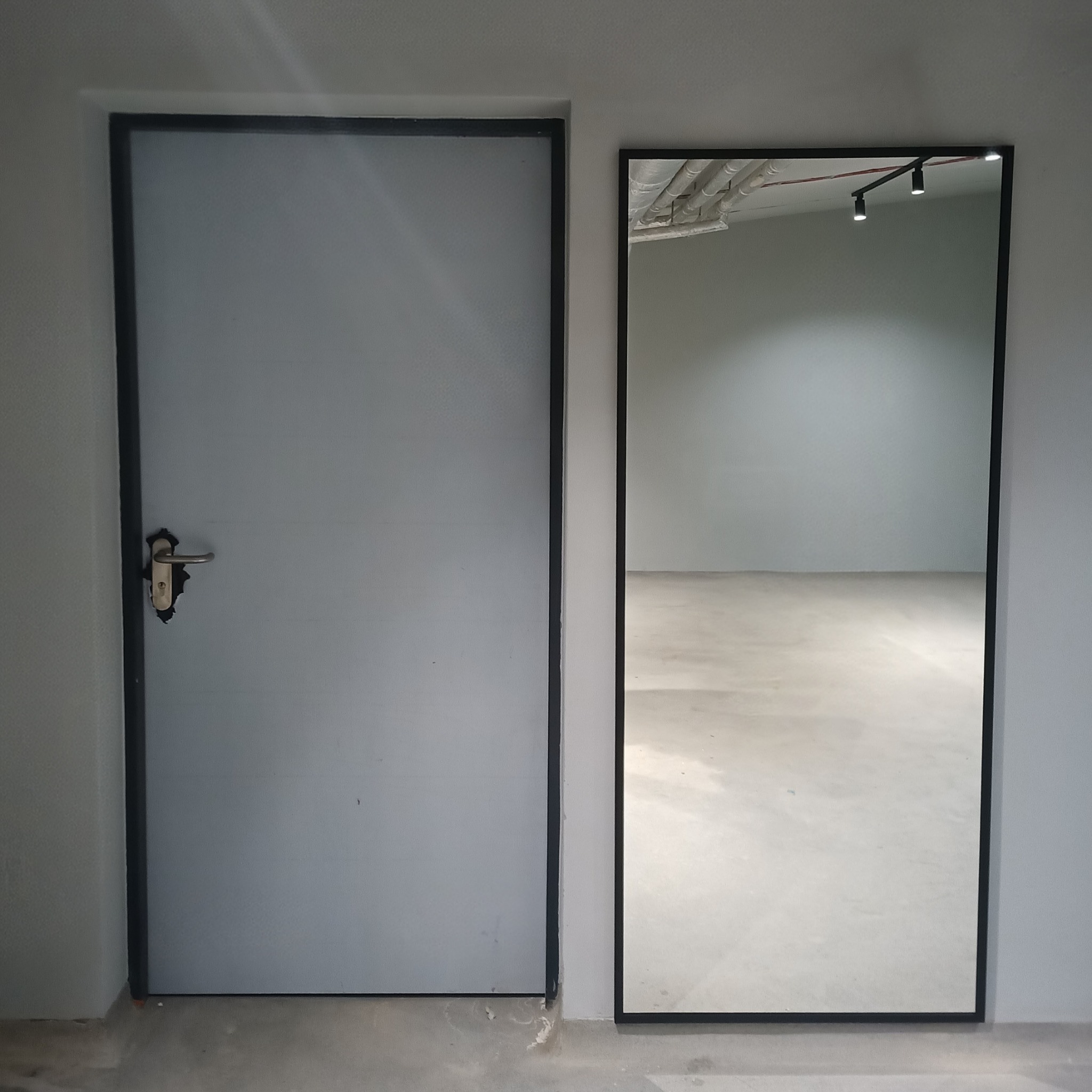 Szara, metalowa futryna drzwi i lustro w czarnej ramie w Skawinie. Widoczne niedoskonałości przy klamce drzwi. W lustrze odbicie sufitu z instalacją.