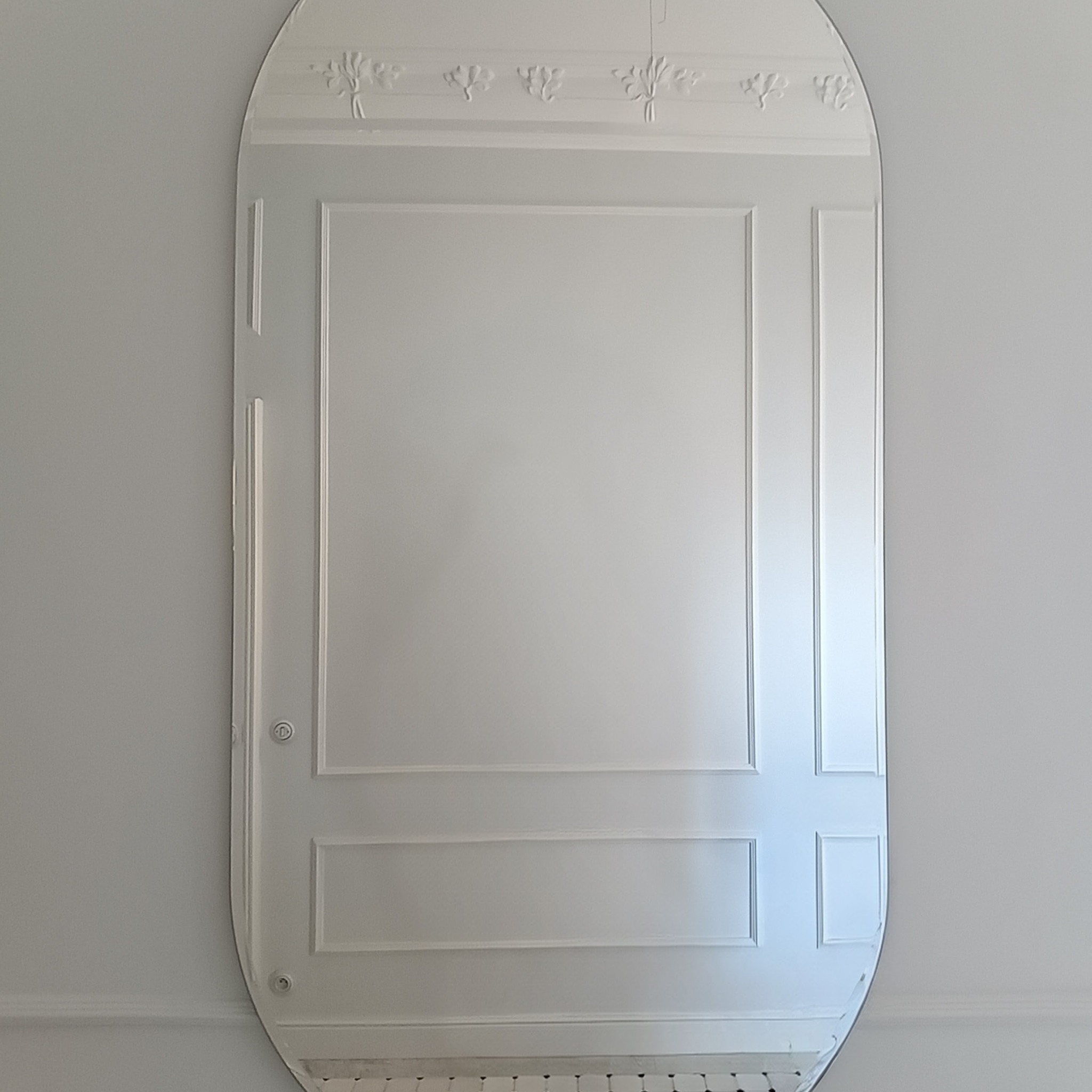 Owalne lustro z dekoracyjnymi listwami i ornamentami, zawieszone na jasnej ścianie. Minimalistyczny design z subtelnymi zdobieniami.