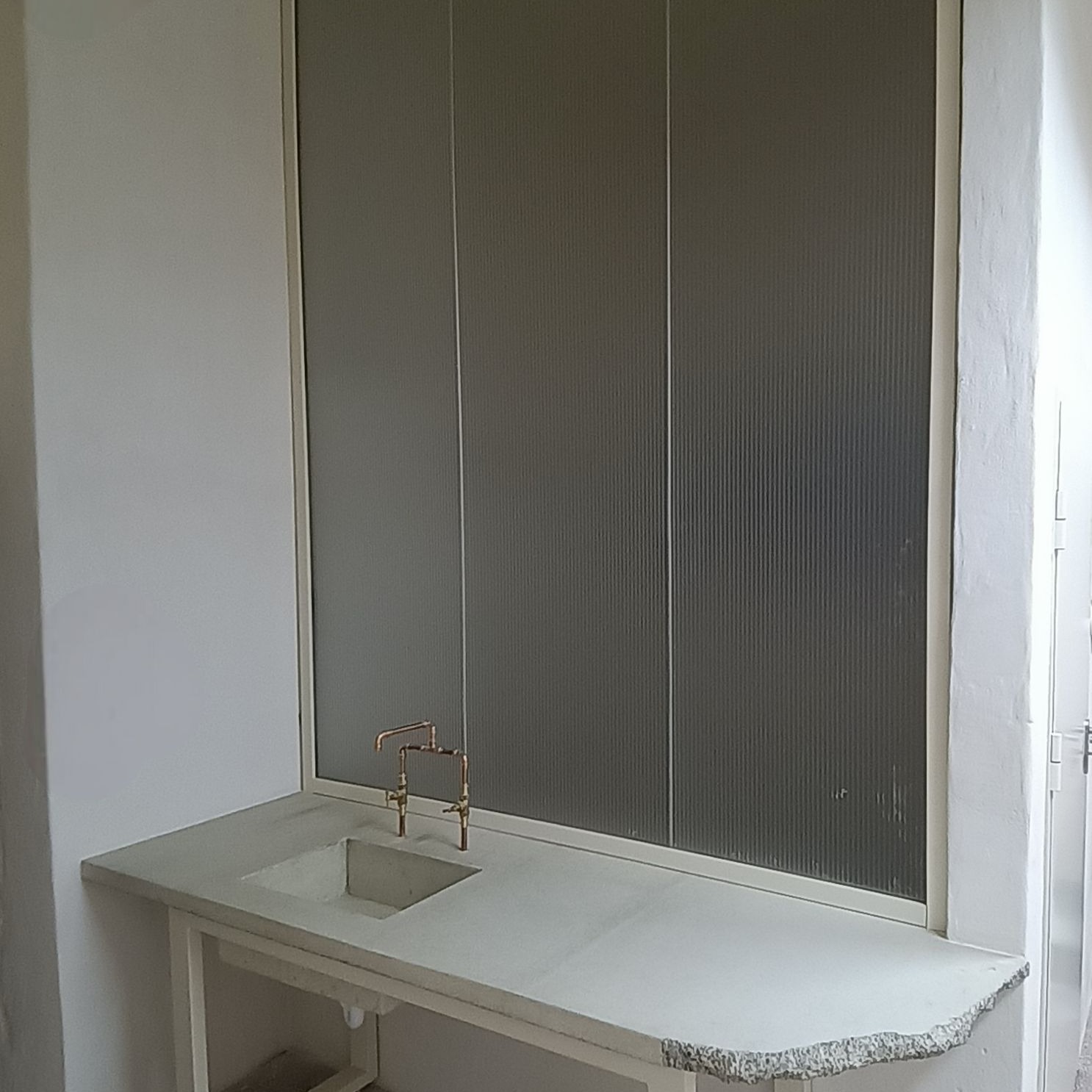 Matowe, ryflowane szkło w łazience nad umywalką z miedzianą armaturą. Minimalistyczny design, jasne wnętrze. Nietypowe rozwiązanie aranżacyjne.