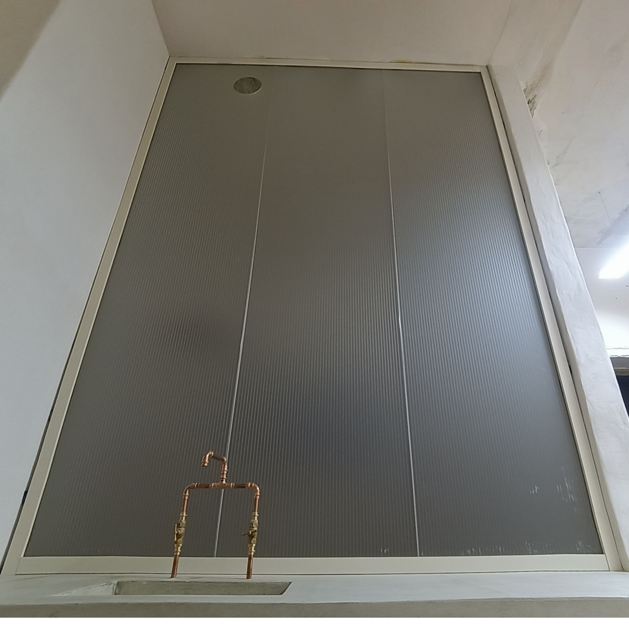 Matowa, szklana ścianka działowa z widoczną instalacją hydrauliczną z miedzi. Ujęcie z dołu podkreśla wysokość konstrukcji w surowym wnętrzu.