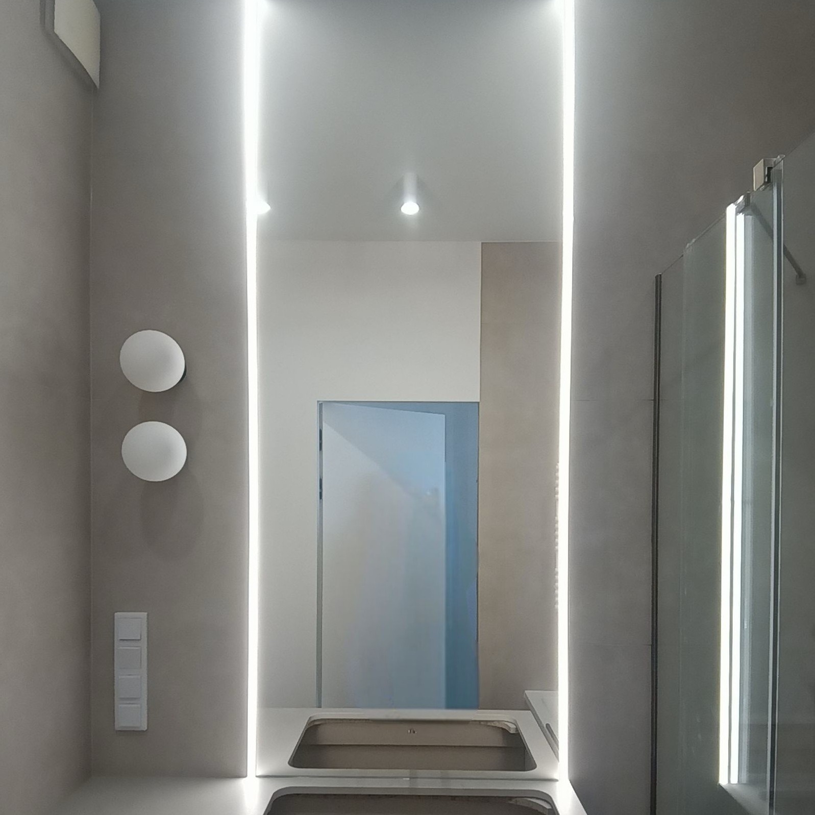 Nowoczesna łazienka z podświetlanym lustrem, szklaną kabiną prysznicową i okrągłymi lampami ściennymi. Minimalistyczny design w odcieniach szarości i bieli.