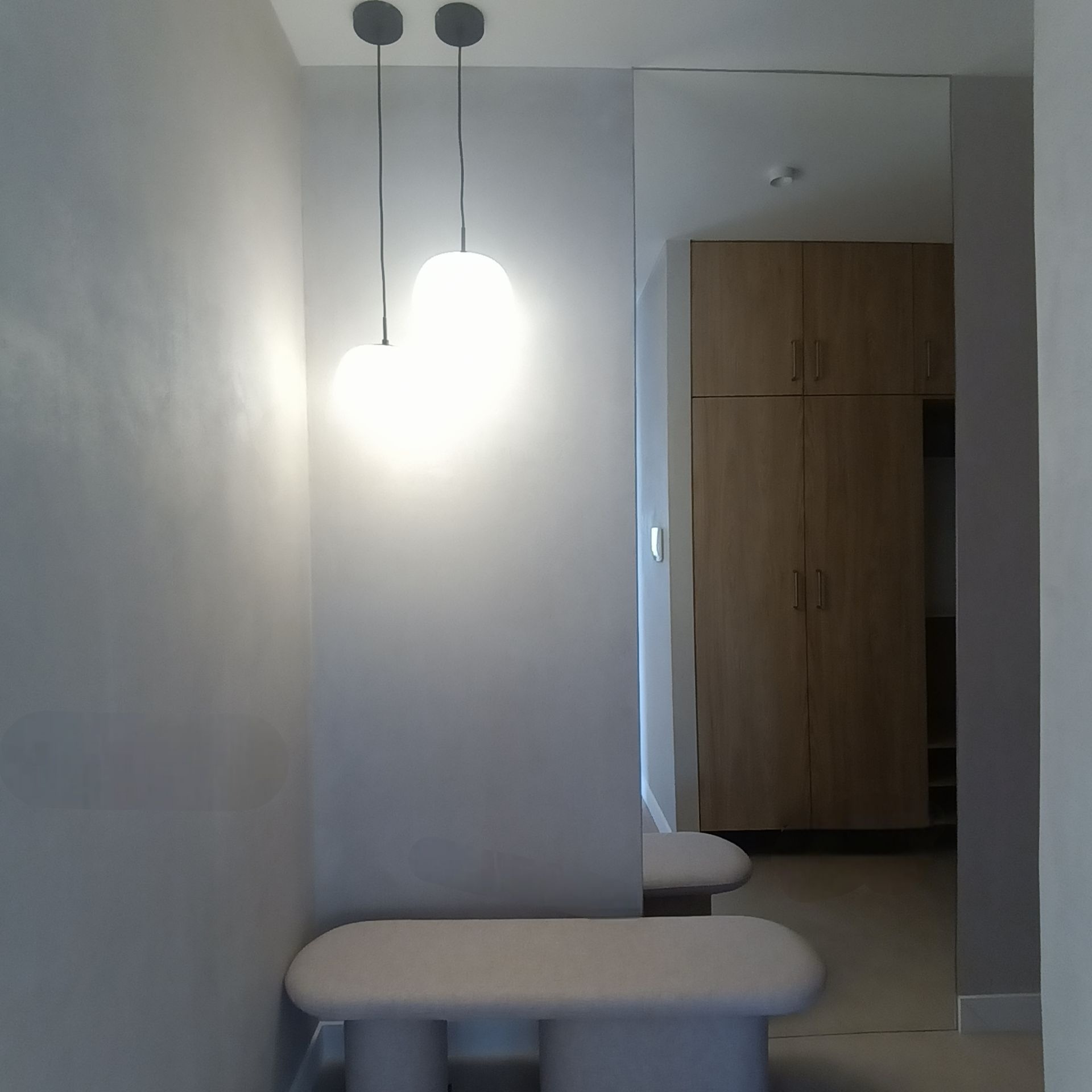 Minimalistyczny przedpokój z lustrem na ścianie, oświetlony dwoma lampami wiszącymi. Widoczna ławka z tapicerowanym siedziskiem i szafa w tle. Jasna kolorystyka.