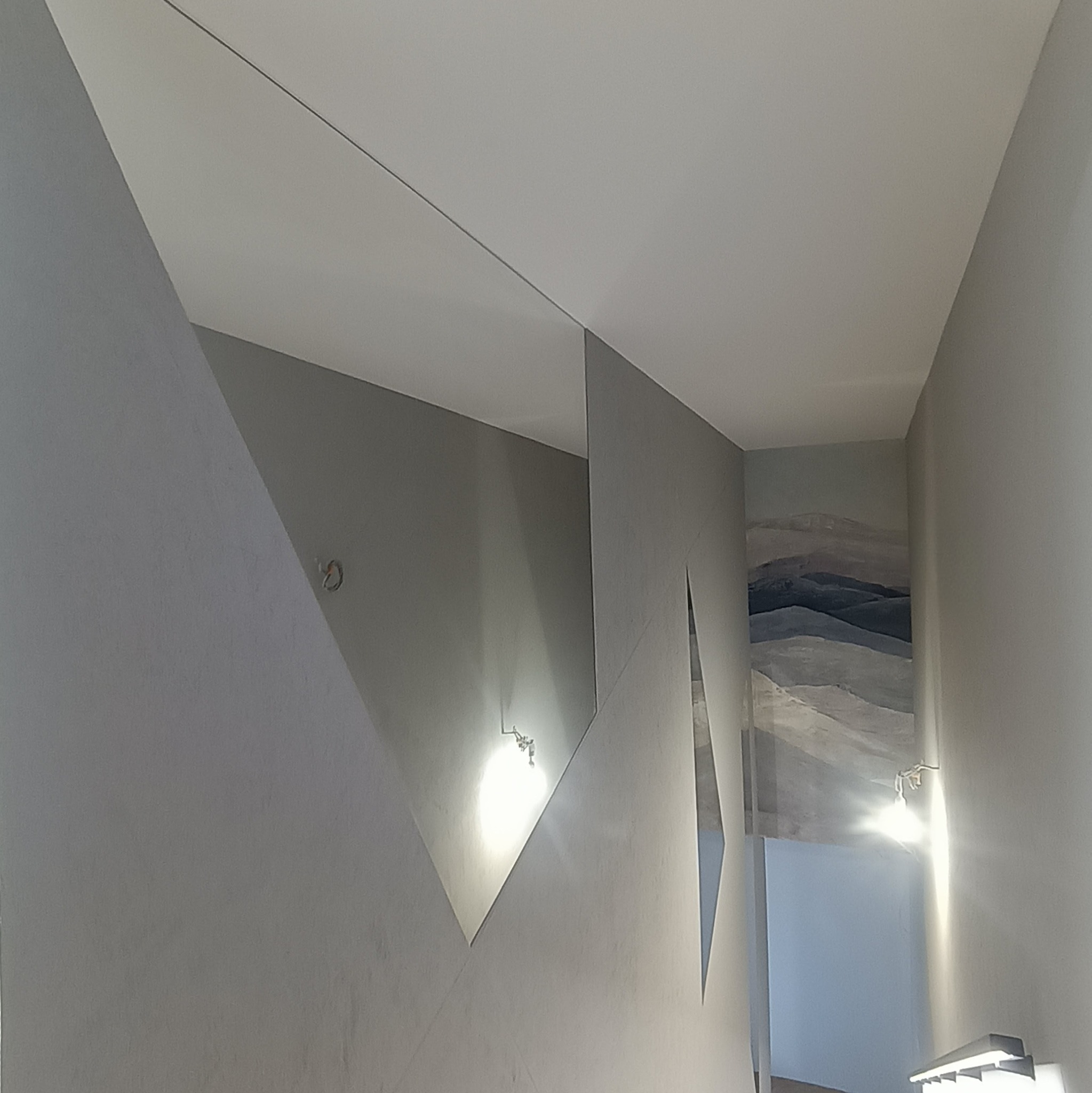 Nowoczesna aranżacja wnętrza z lustrami o geometrycznych kształtach na ścianie, oświetlonych lampami. Widok z dołu, szara kolorystyka.