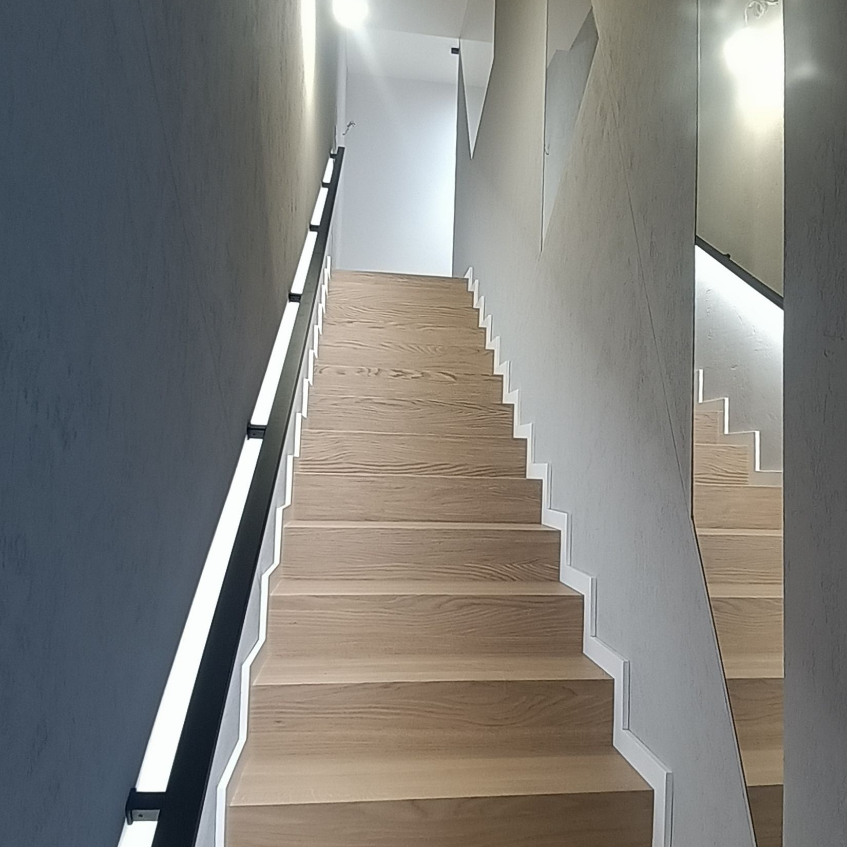 Nowoczesne schody drewniane z podświetlaną balustradą i lustrem na ścianie. Minimalistyczny design, jasne drewno, szara ściana. Ujęcie od dołu, podkreślające geometrię.