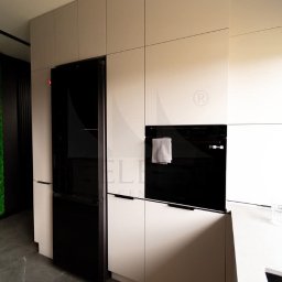Stolarnia Meble - Biała, nowoczesna zabudowa kuchenna z czarnym piekarnikiem i lodówką side-by-side, na ścianie zielony panel z mchu.