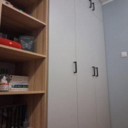 Stolarnia Meble - Szara szafa wnękowa z czarnymi, prostokątnymi uchwytami, sąsiadująca z drewnianym regałem wypełnionym książkami i ozdobami.