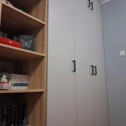 Szara szafa wnękowa z czarnymi, prostokątnymi uchwytami, sąsiadująca z drewnianym regałem wypełnionym książkami i ozdobami.