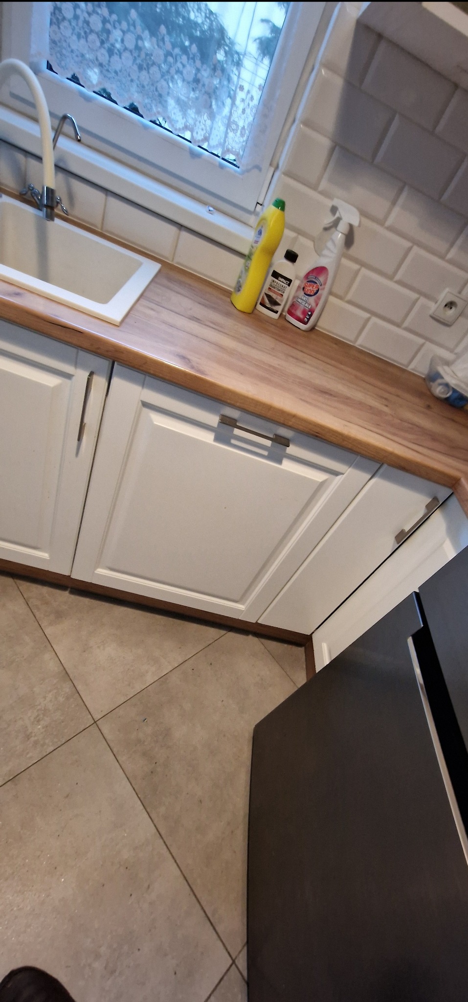 Fragment blatu kuchennego z białym zlewozmywakiem i baterią, widoczne środki czystości oraz białe fronty szafek z poziomymi uchwytami, płytki ceramiczne na ścianie i fragment lodówki w tle.