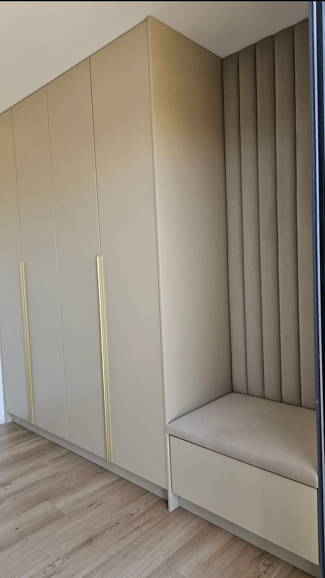 Beżowa szafa wnękowa z długimi złotymi uchwytami, połączona z tapicerowanym siedziskiem w pionowe pasy, na drewnopodobnej podłodze.
