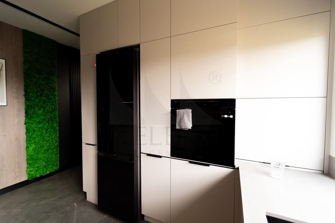 Biała, nowoczesna zabudowa kuchenna z czarnym piekarnikiem i lodówką side-by-side, na ścianie zielony panel z mchu.