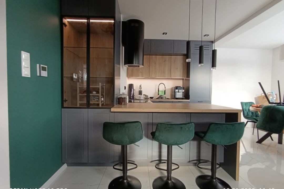 Nowoczesna kuchnia z szarymi szafkami, drewnianymi akcentami, czarnym okapem i barkiem z zielonymi hokerami, oświetlona wiszącymi lampami.