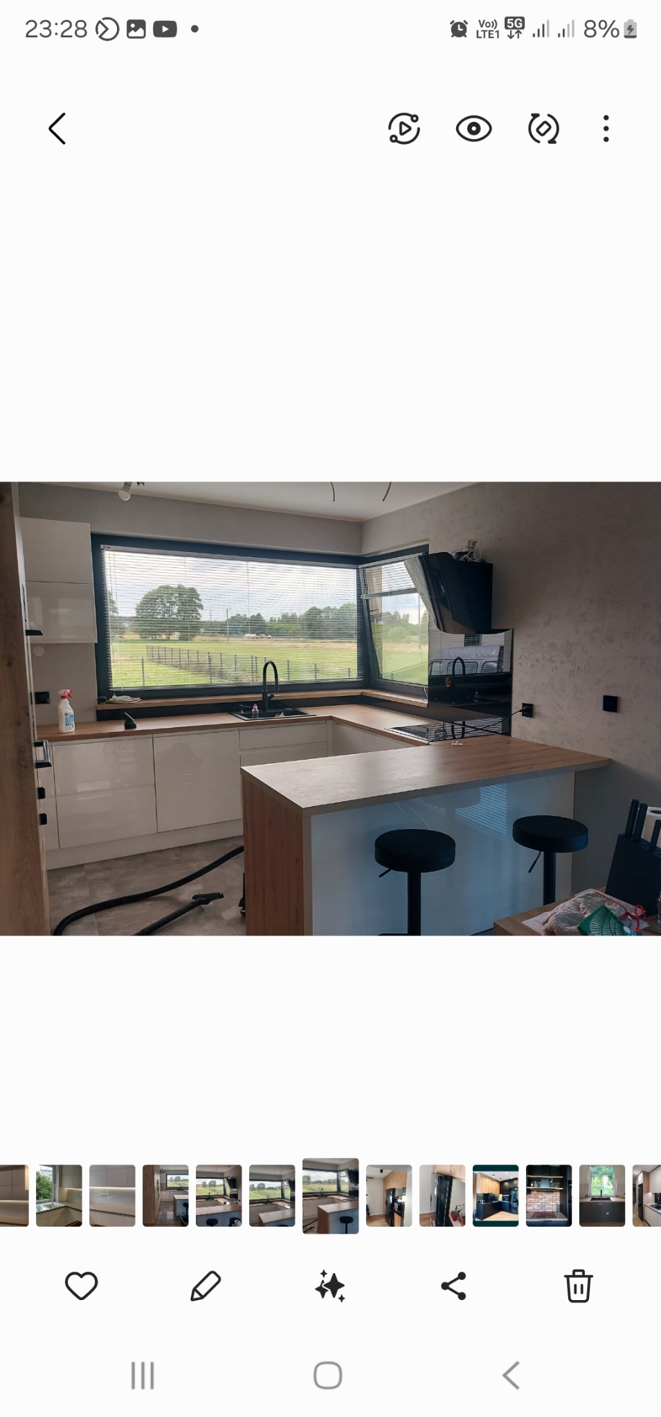 Nowoczesna kuchnia z białymi szafkami i drewnianym blatem, wyspą z hokerami oraz dużym oknem z widokiem na zielony krajobraz, czyszczona odkurzaczem.