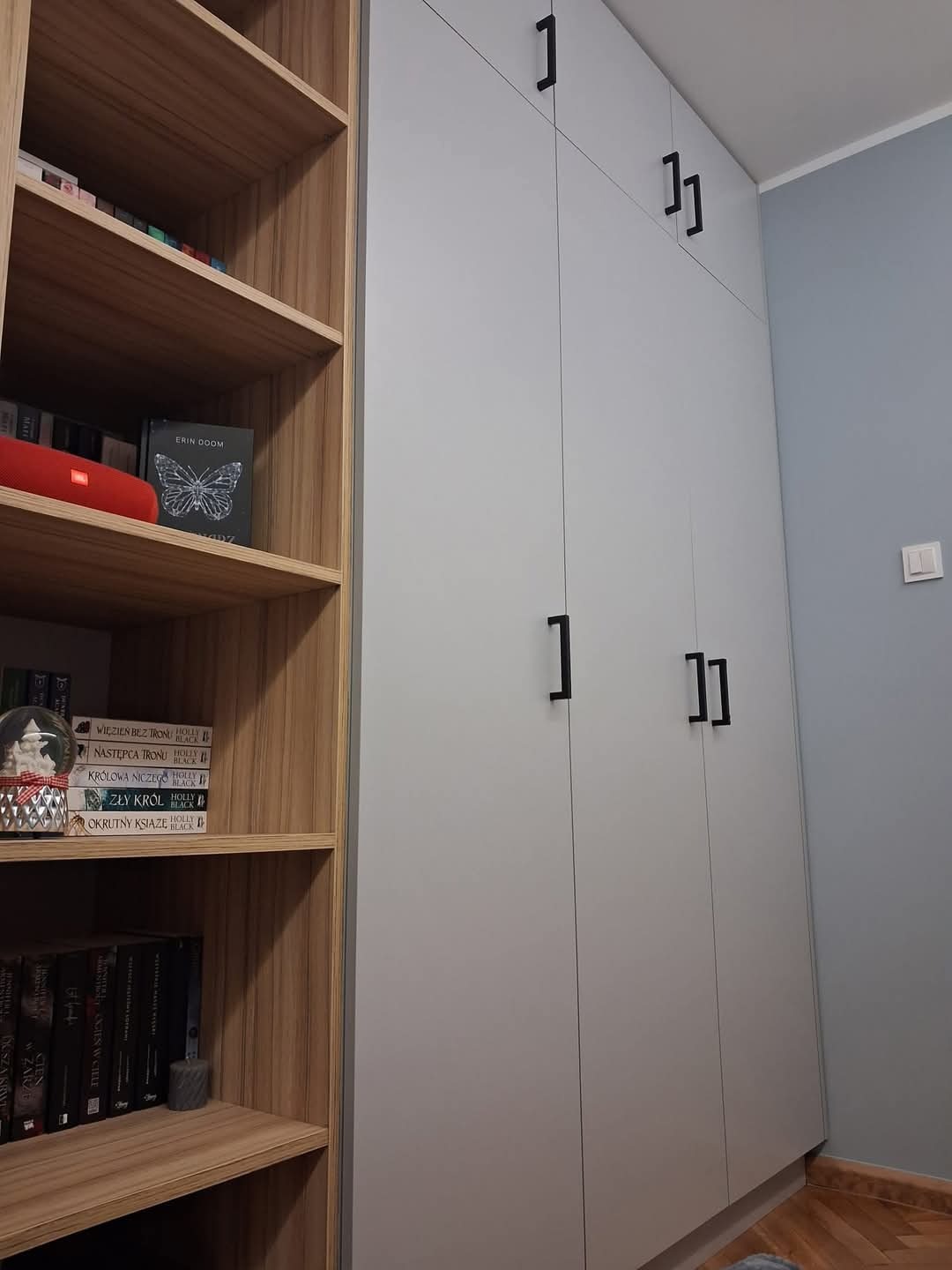 Szara szafa wnękowa z czarnymi, prostokątnymi uchwytami, sąsiadująca z drewnianym regałem wypełnionym książkami i ozdobami.