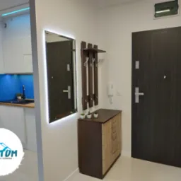 Nowoczesny przedpokój z podświetlanym lustrem, komodą z wieszakami i ciemnymi drzwiami wejściowymi. W tle widoczna kuchnia z niebieskim panelem szklanym.