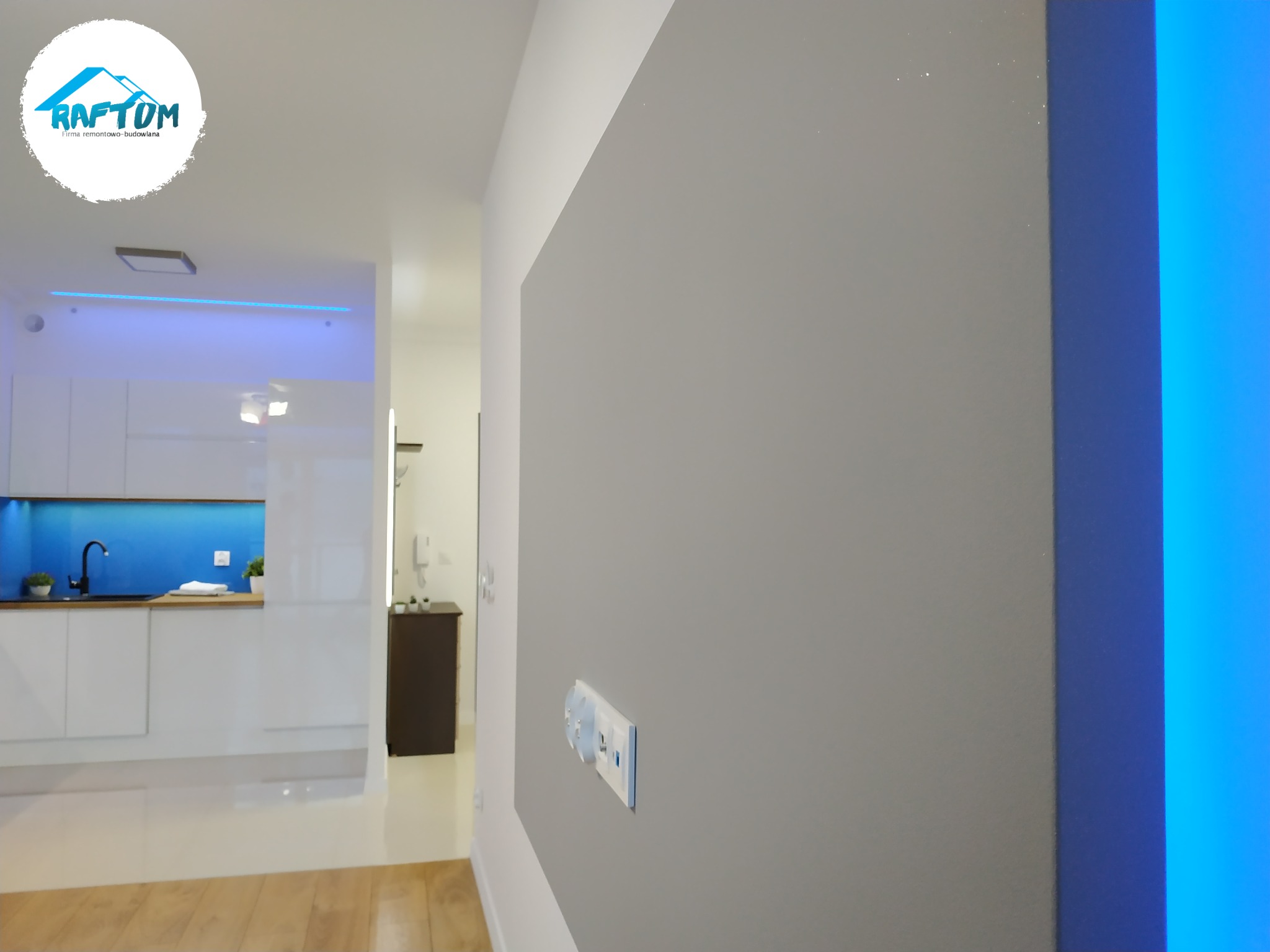 Szara ściana z gładzią i widocznymi gniazdkami w nowocześnie wykończonym mieszkaniu, z widokiem na białą kuchnię z niebieskim oświetleniem LED w tle. W lewym górnym rogu widoczne logo firmy...