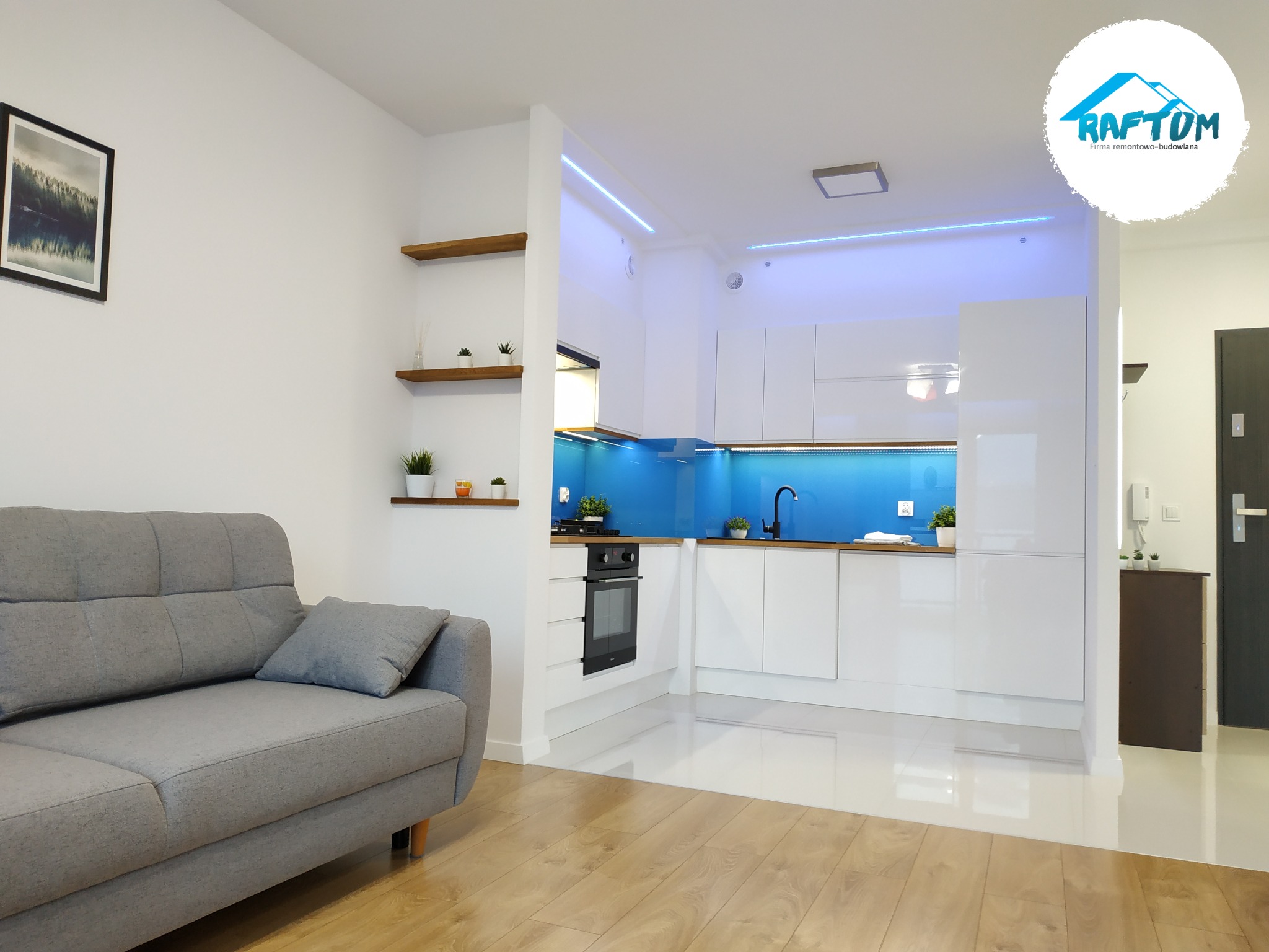 Nowoczesne wnętrze mieszkania z szarą sofą i białą kuchnią z niebieskim panelem, drewniana podłoga, oświetlenie LED pod szafkami, obraz na ścianie oraz logo firmy remontowej w prawym górnym rogu.