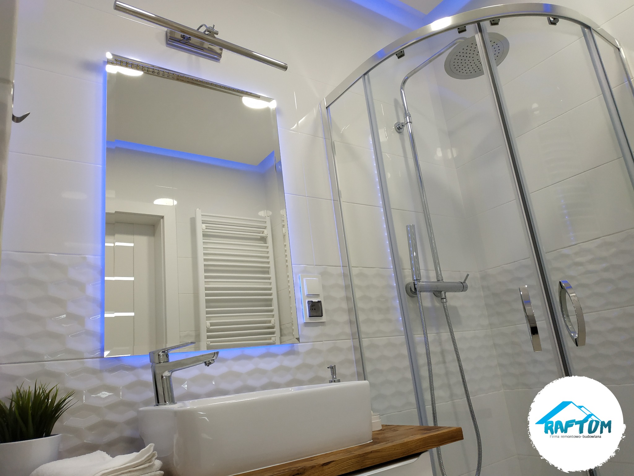 Nowoczesna łazienka z białą armaturą, kabiną prysznicową z deszczownicą, umywalką nablatową na drewnianym blacie i lustrem podświetlanym niebieskim światłem, widoczne logo firmy.