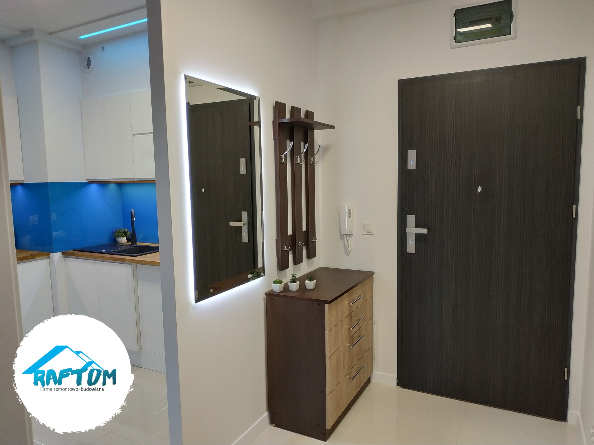 Nowoczesny przedpokój z podświetlanym lustrem, komodą z wieszakami i ciemnymi drzwiami wejściowymi. W tle widoczna kuchnia z niebieskim panelem szklanym.