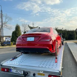 Czerwone sportowe auto na srebrnej lawecie, zabezpieczone czerwonymi pasami. Widok z tyłu, tablica rejestracyjna widoczna. Błękitne niebo w tle.