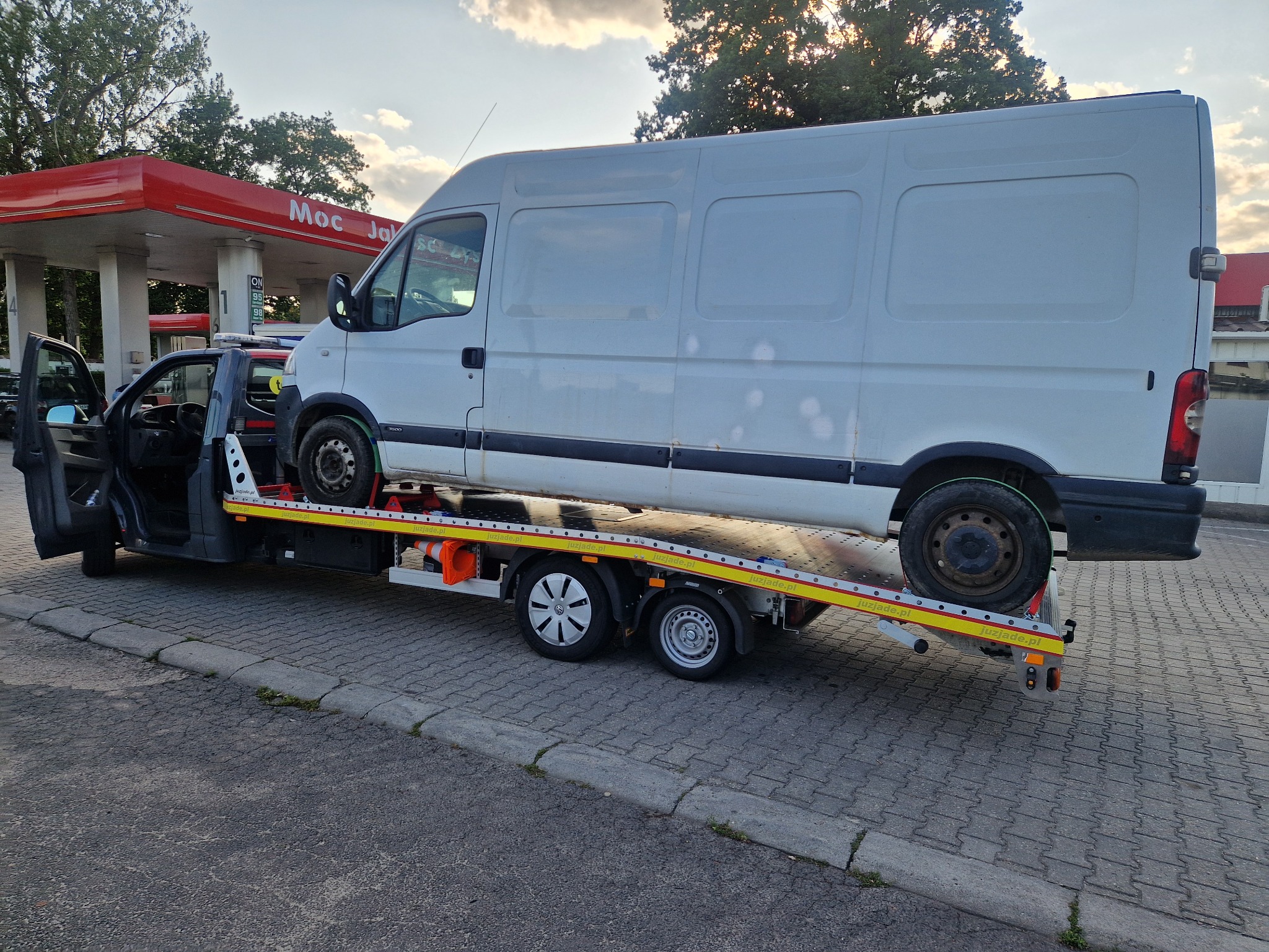 Laweta z białym busem na platformie, na tle stacji benzynowej. Widoczne oznaczenia lawety i częściowo wnętrze kabiny. Ujęcie z boku, w dziennym świetle.