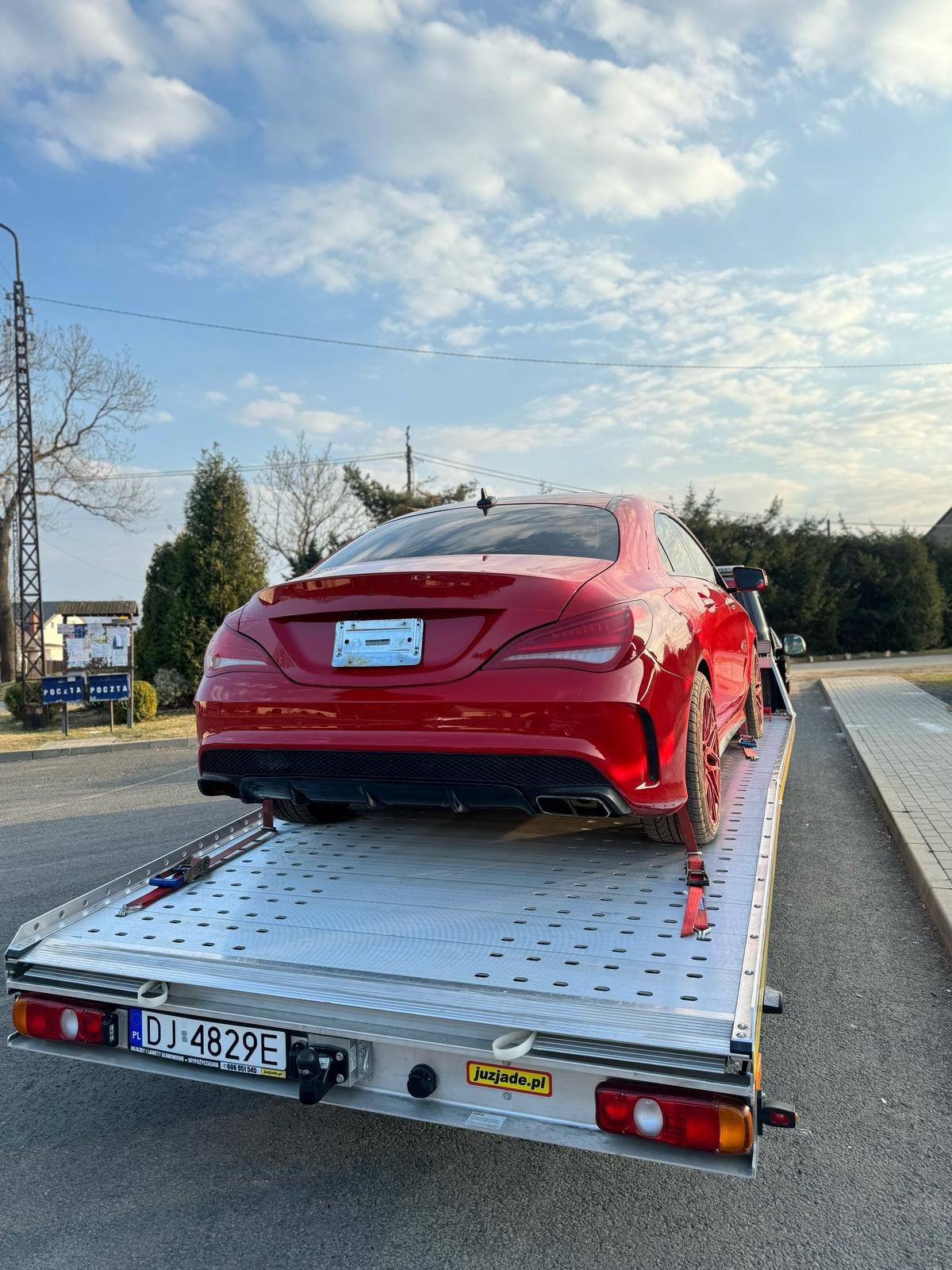 Czerwone sportowe auto na srebrnej lawecie, zabezpieczone czerwonymi pasami. Widok z tyłu, tablica rejestracyjna widoczna. Błękitne niebo w tle.