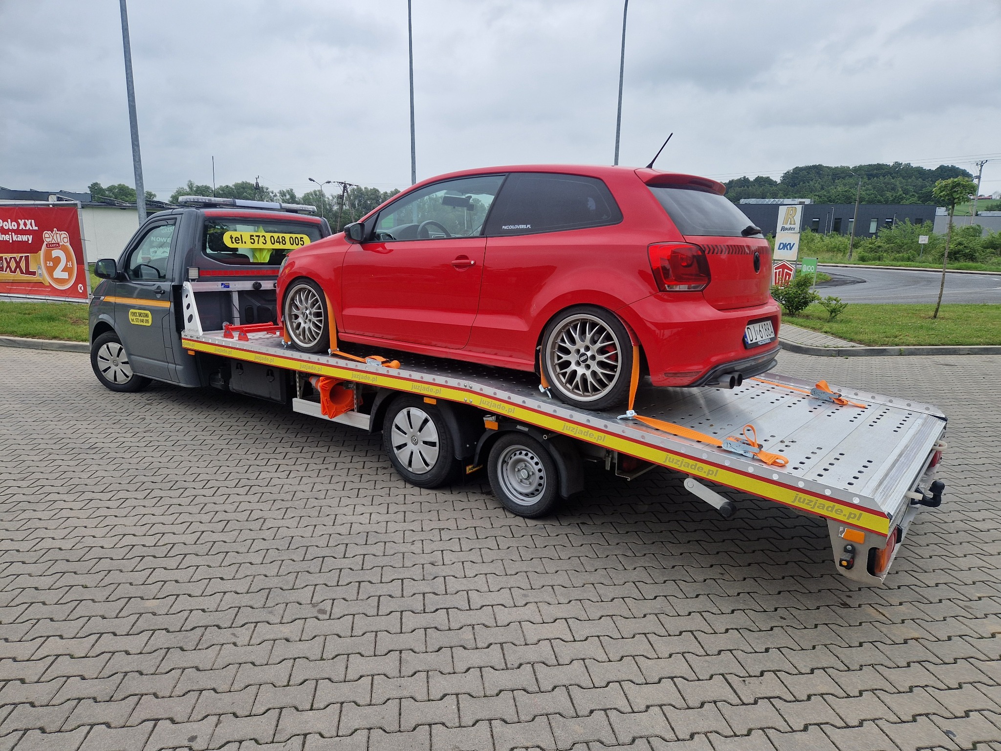 Czerwony Volkswagen Polo na srebrnej lawecie z pomarańczowymi pasami mocującymi, zaparkowanej na kostce brukowej. Widoczny numer telefonu na lawecie.