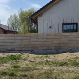 Wznoszenie ogrodzenia z beżowych, modułowych bloczków betonowych, z wystającymi prętami zbrojeniowymi, na tle szarego budynku w trakcie budowy i drewnianego domku gospodarczego.
