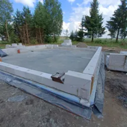 Wznoszenie ścian fundamentowych z bloczków betonowych na przygotowanej płycie, widoczne narzędzia murarskie i rozciągnięte sznurki, w tle zieleń i błękitne niebo.