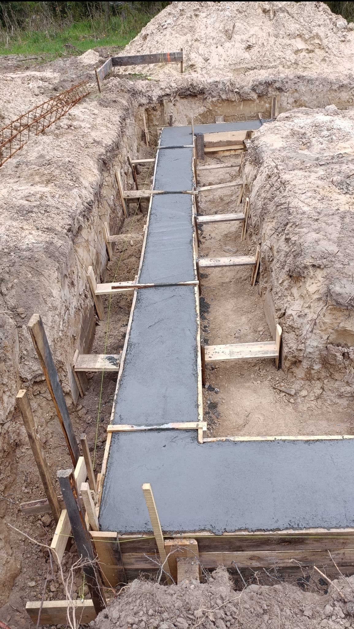 Wylany beton w szalunku fundamentowym, widoczny wykop z ziemią i zbrojenie na obrzeżach.