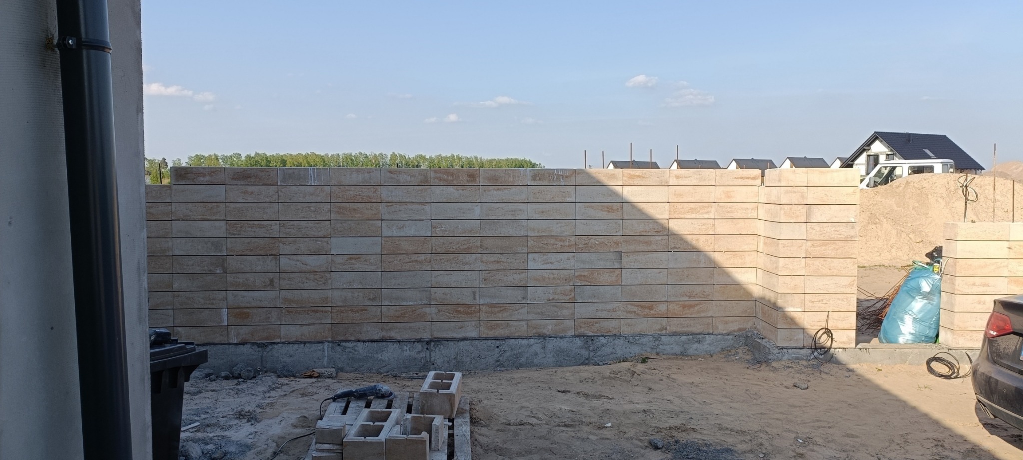 Wznoszona ściana z bloczków z betonu architektonicznego, widoczne narzędzia, piasek i fragment samochodu, w tle zabudowa jednorodzinna.