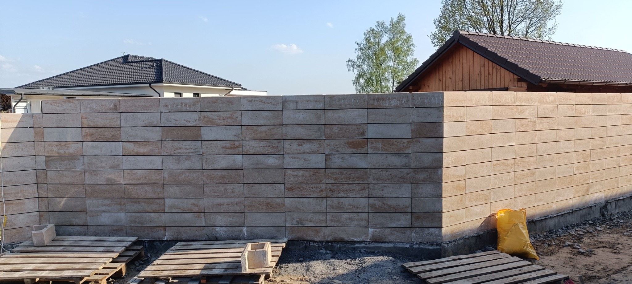 Wzniesiony fragment ogrodzenia z beżowych bloczków betonowych z widocznymi dwoma domami w tle, jeden z ciemną dachówką, drugi z drewnianą elewacją.