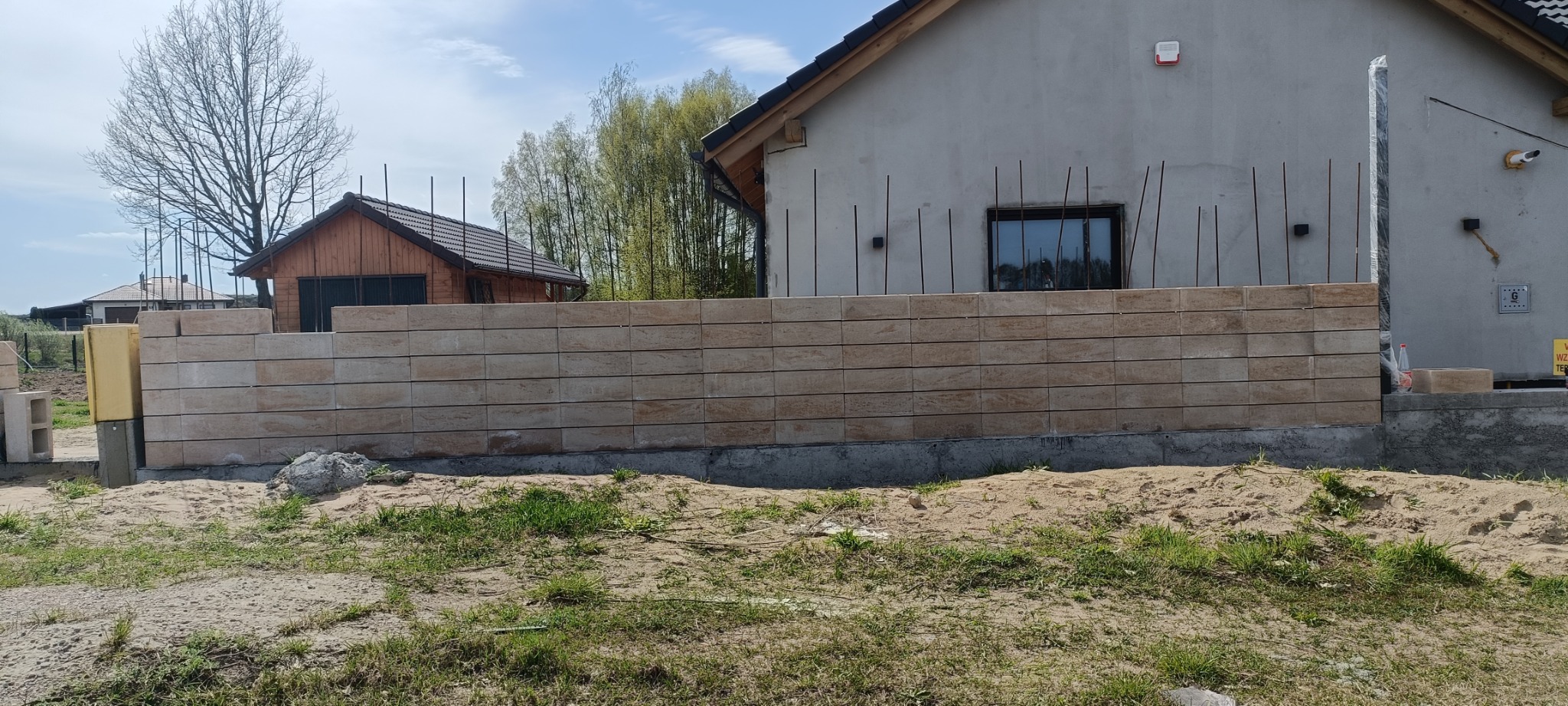 Wznoszenie ogrodzenia z beżowych, modułowych bloczków betonowych, z wystającymi prętami zbrojeniowymi, na tle szarego budynku w trakcie budowy i drewnianego domku gospodarczego.