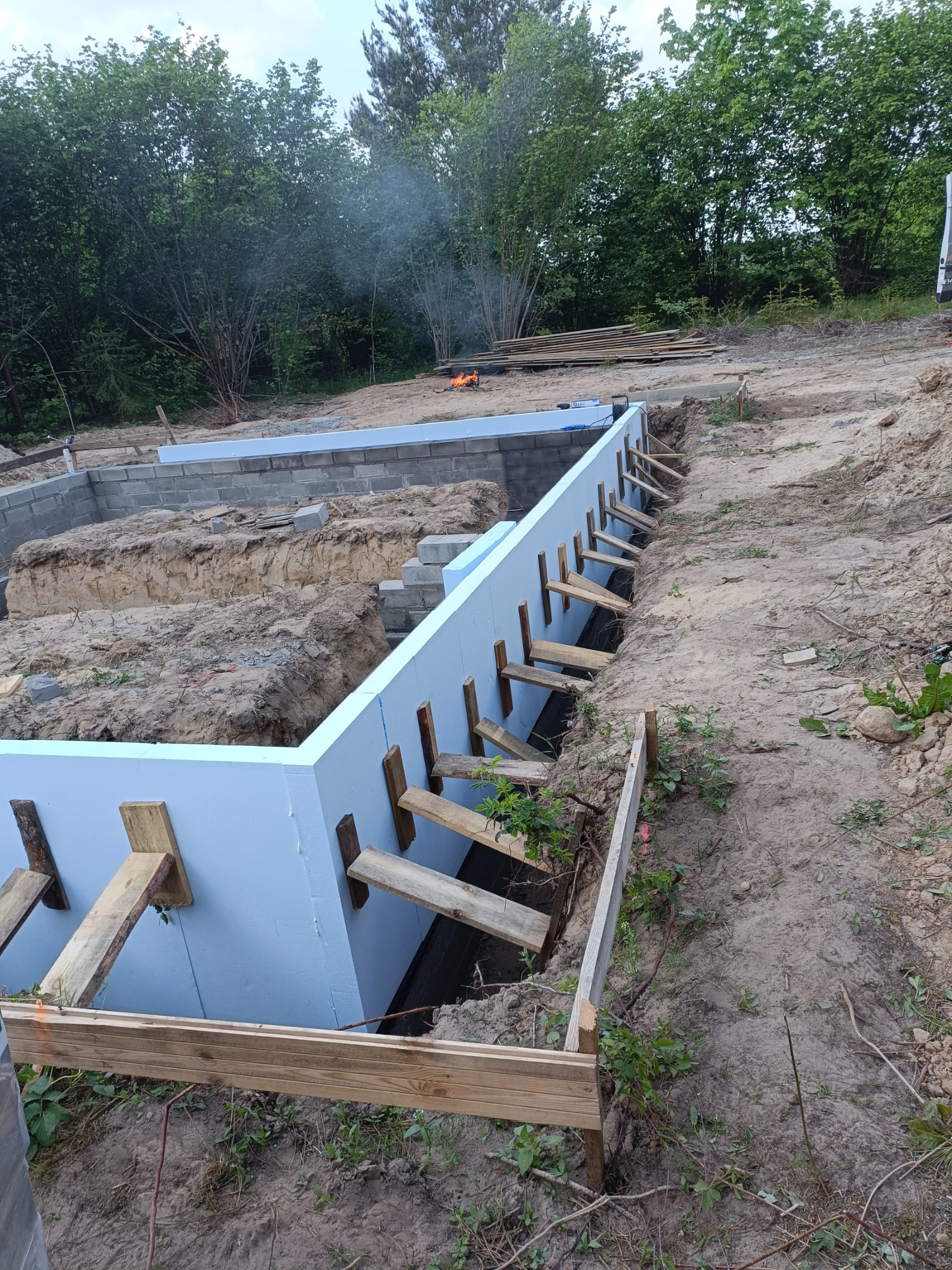 Izolacja fundamentów budynku z użyciem niebieskiego styropianu, zabezpieczona drewnianymi stemplami, widok z góry na wykop z elementami konstrukcji murowanej w tle, na ziemi widoczne rośliny...