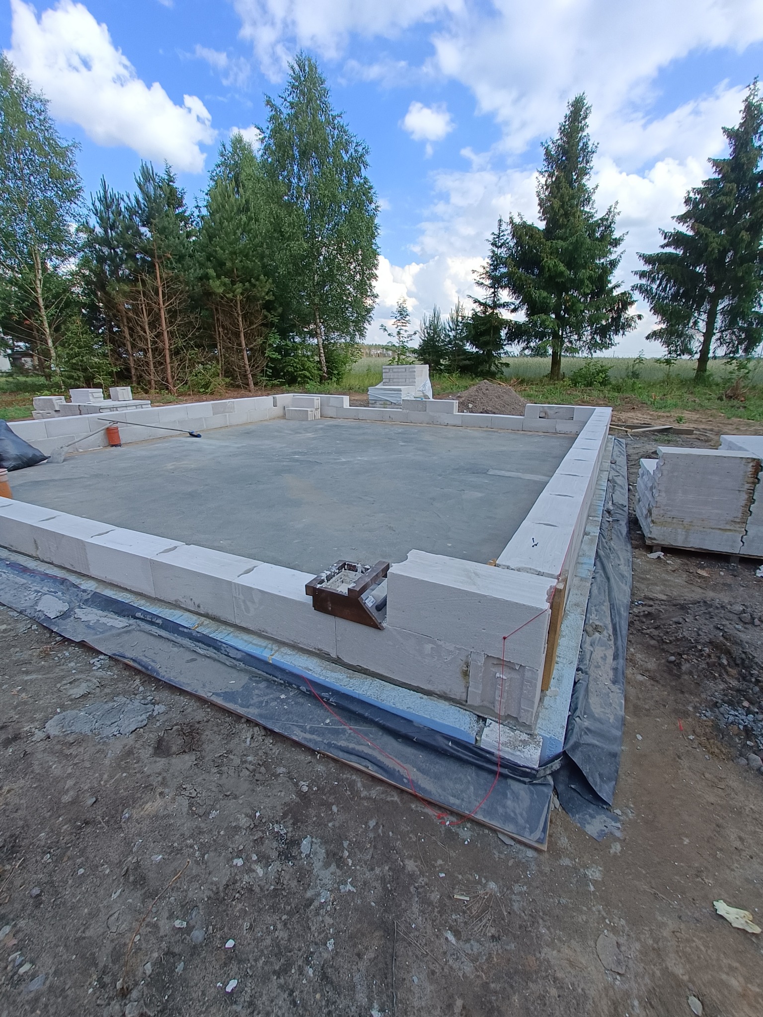 Wznoszenie ścian fundamentowych z bloczków betonowych na przygotowanej płycie, widoczne narzędzia murarskie i rozciągnięte sznurki, w tle zieleń i błękitne niebo.