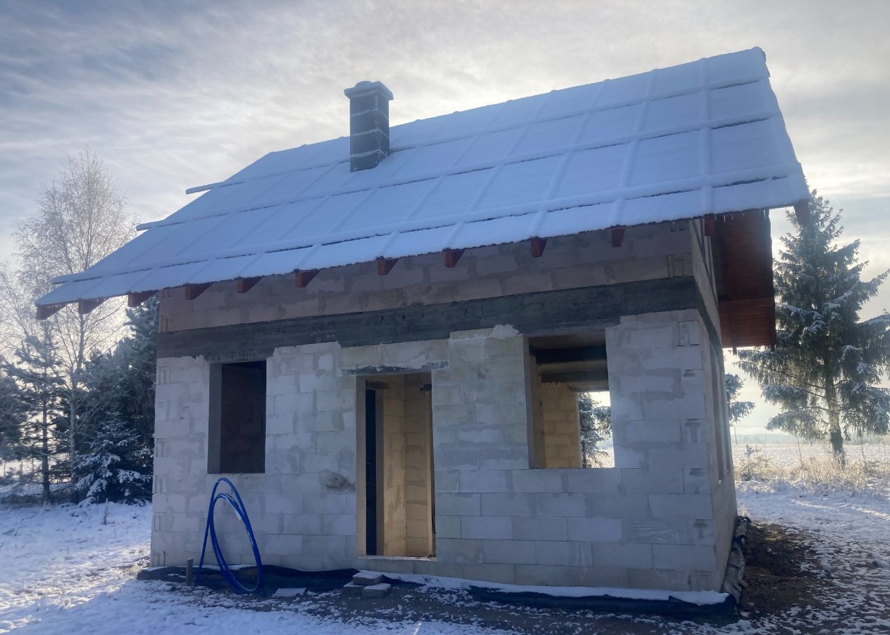 Niedokończony dom z białego bloczka, z oknami i drzwiami bez osadzenia, pokryty śniegiem, otoczony zimowym krajobrazem z drzewami i polem.