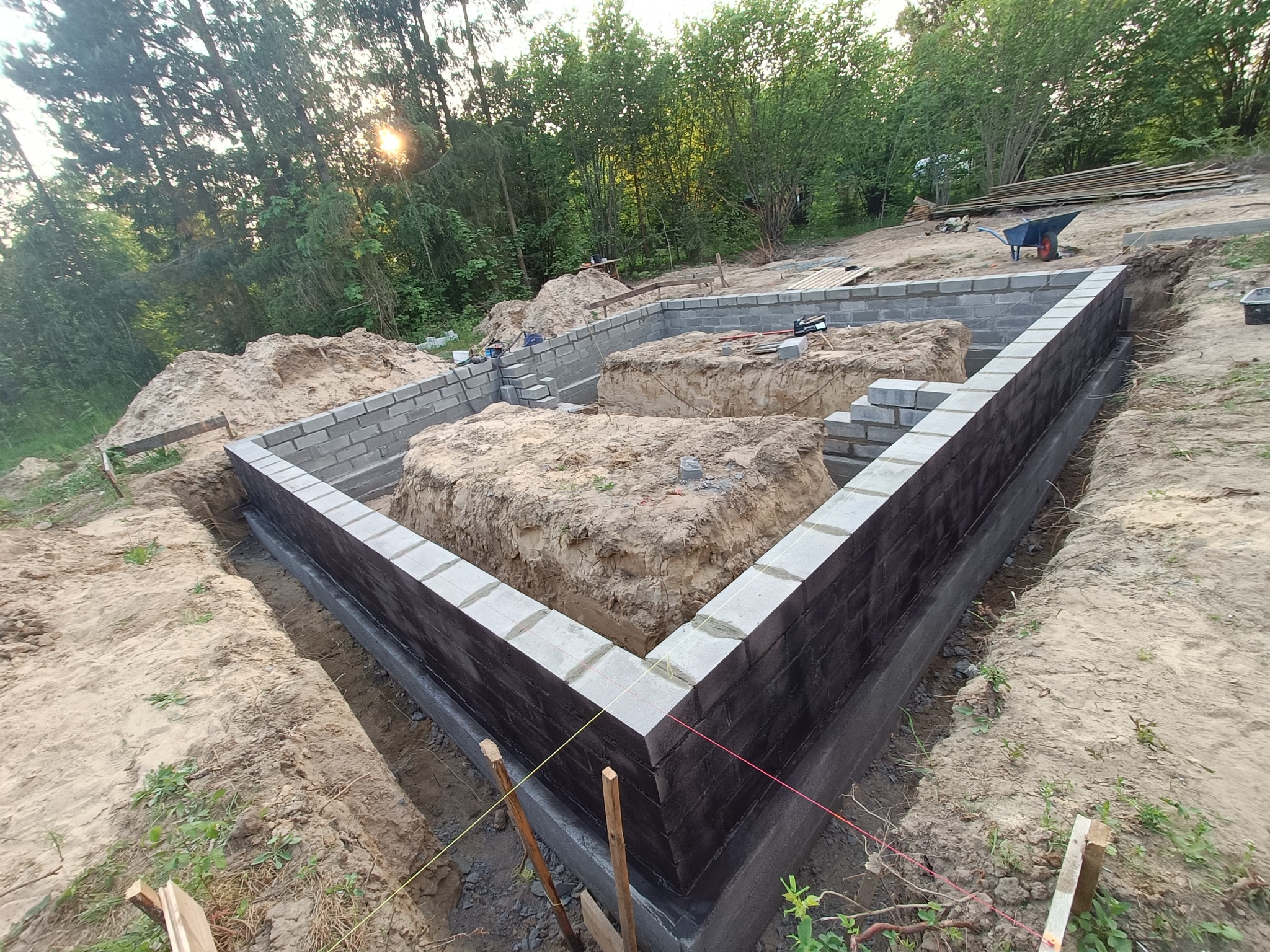 Wykop pod fundamenty z częściowo wzniesionymi ścianami z szarych bloczków betonowych, izolowanymi od zewnątrz czarną masą bitumiczną, na tle drzew i zachodzącego słońca.