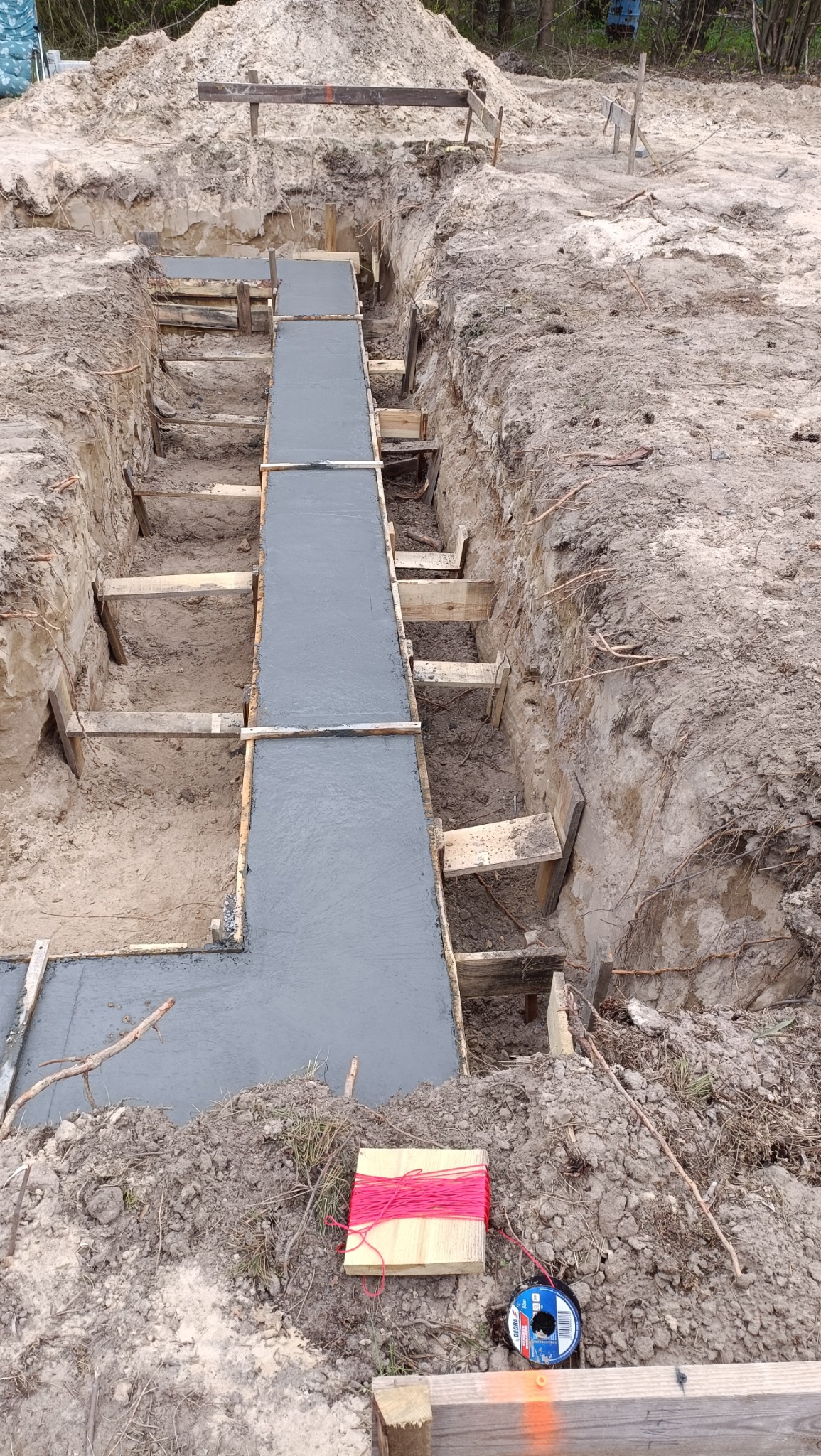 Wykop pod fundamenty zalany świeżym betonem, widoczne drewniane szalunki i poziomica sznurkowa na ziemi.