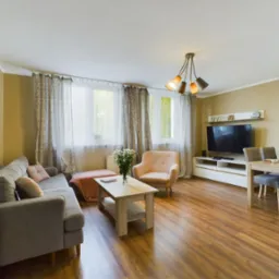 Jasne, przestronne wnętrze salonu z drewnianą podłogą, szarą sofą, beżowym fotelem, stolikiem kawowym z kwiatami, stołem jadalnym z szarymi krzesłami i telewizorem na białej komodzie.