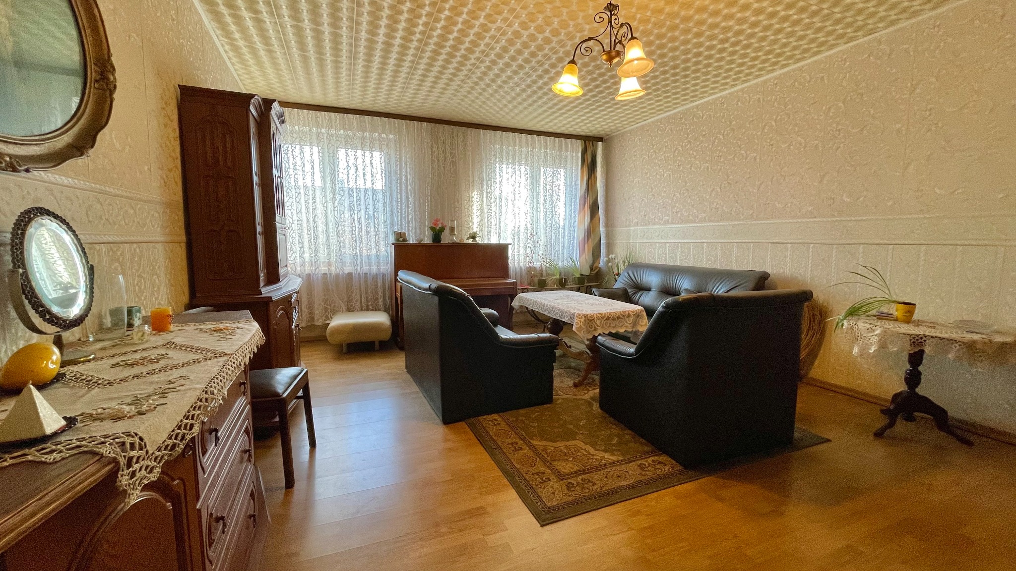 Umeblowany pokój dzienny w stylu retro z ciemnymi skórzanymi fotelami, sofą, pianinem pod oknem i ozdobną komodą z lustrami, oświetlony lampą z żółtymi kloszami.