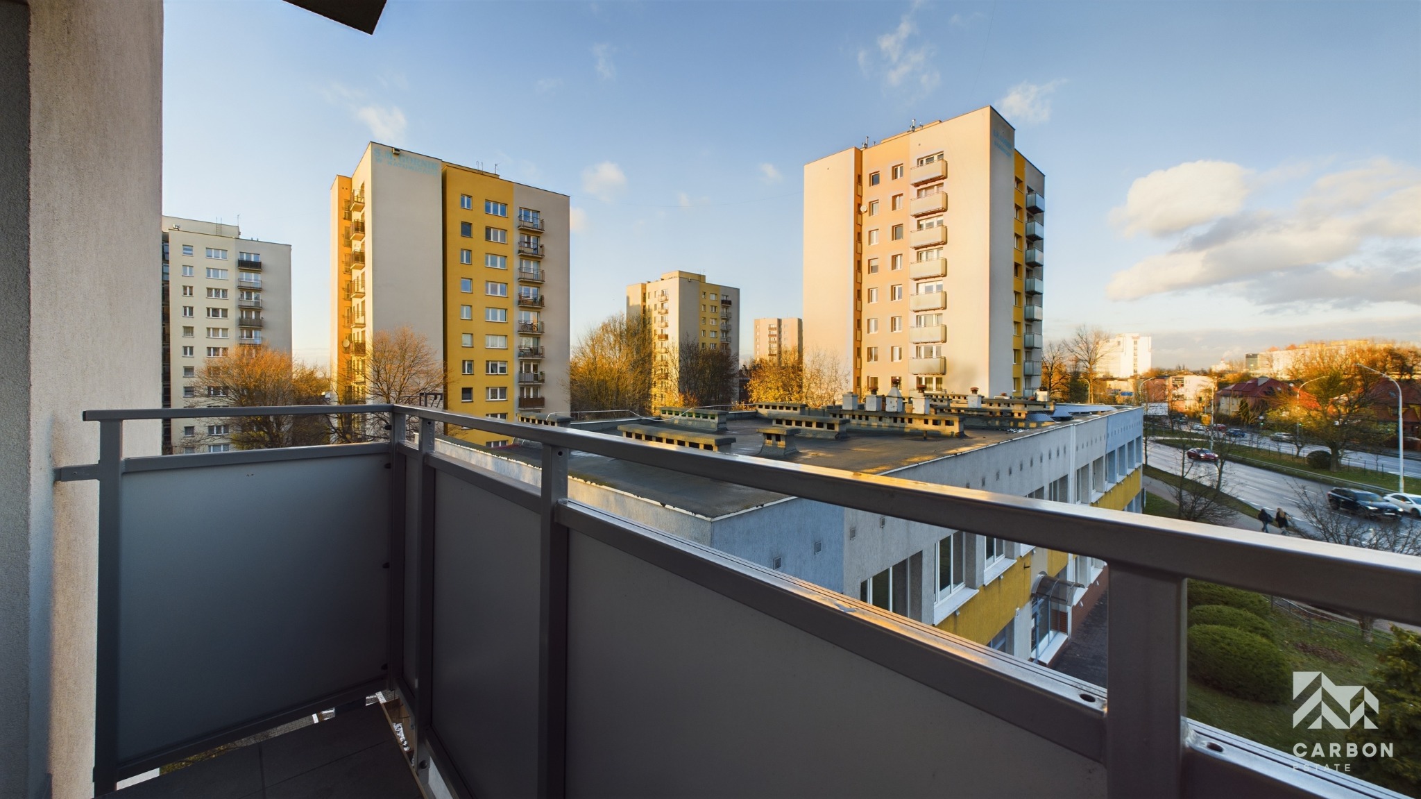 Widok z balkonu w Katowicach na osiedle bloków mieszkalnych w jesiennym słońcu, z widoczną metalową barierką i fragmentem płyty balkonowej.