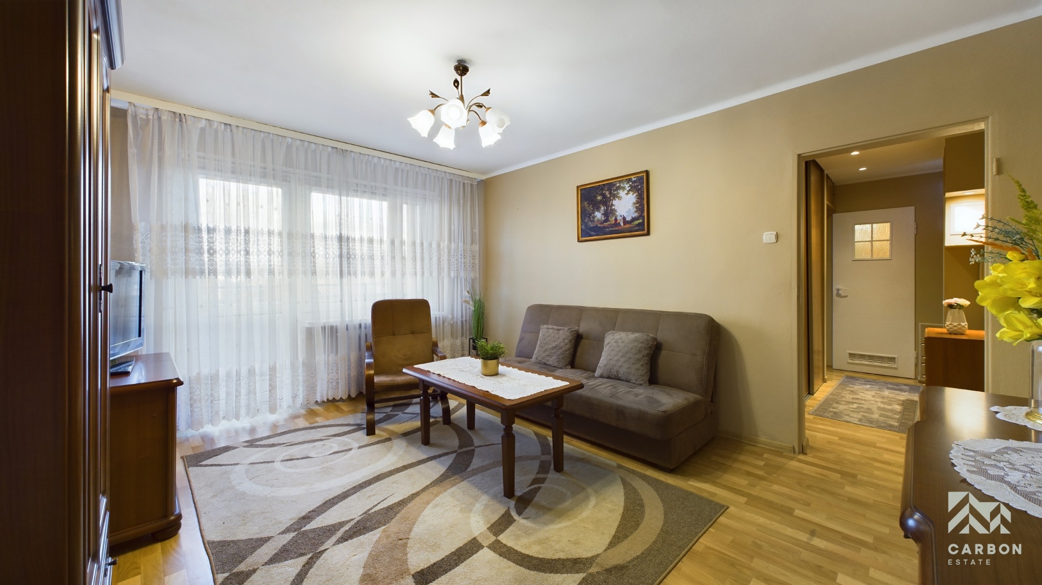 Jasny salon z brązową sofą, fotelem i stolikiem kawowym na wzorzystym dywanie, widok na okno z białymi zasłonami i drzwi do innego pomieszczenia, oświetlenie sufitowe.