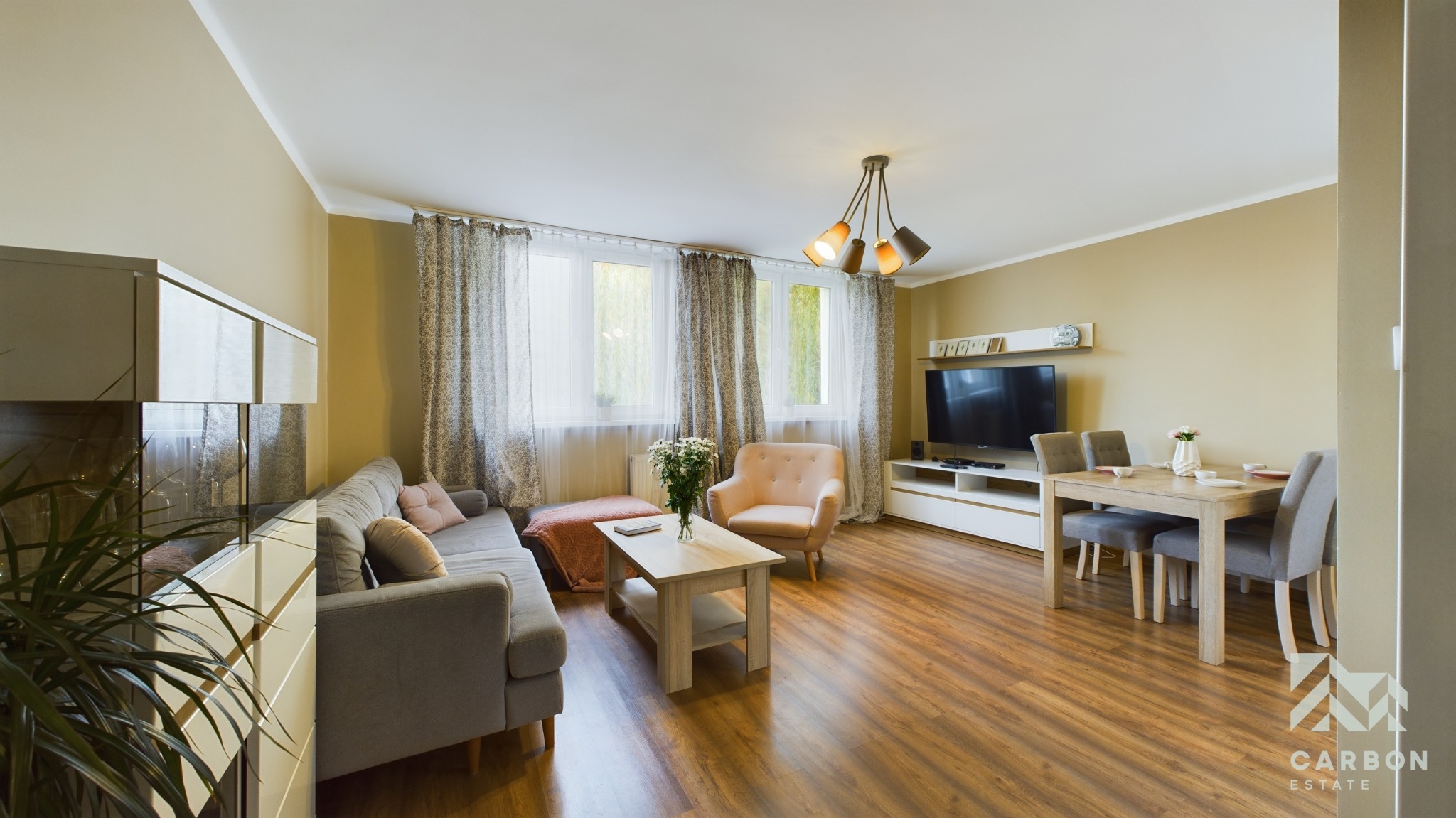 Jasne, przestronne wnętrze salonu z drewnianą podłogą, szarą sofą, beżowym fotelem, stolikiem kawowym z kwiatami, stołem jadalnym z szarymi krzesłami i telewizorem na białej komodzie.
